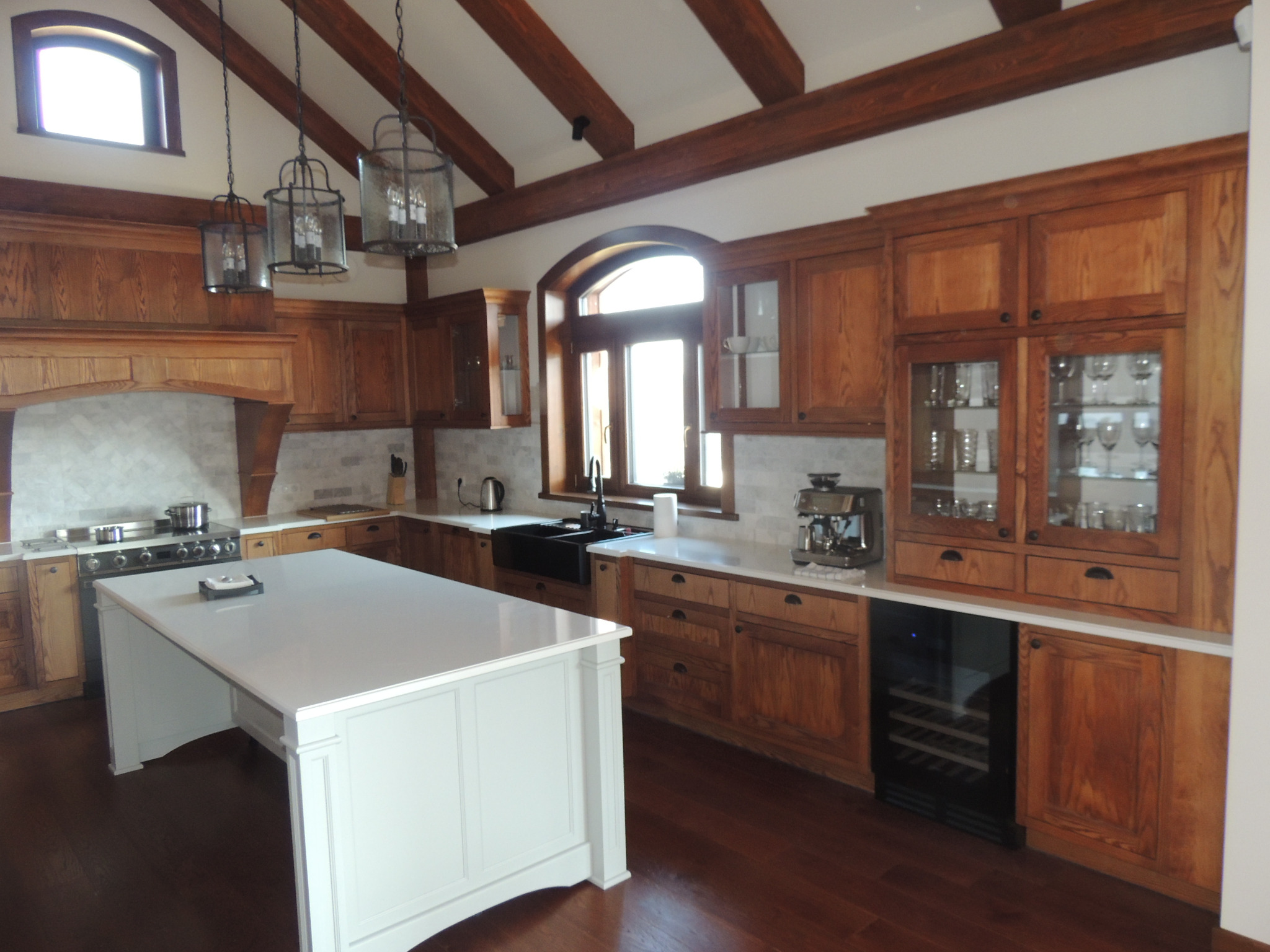 Rustykalna kuchnia z drewnianymi szafkami, białym blatem wyspy i dekoracyjnymi lampami wiszącymi, widok na okno łukowe i drewniane belki stropowe.