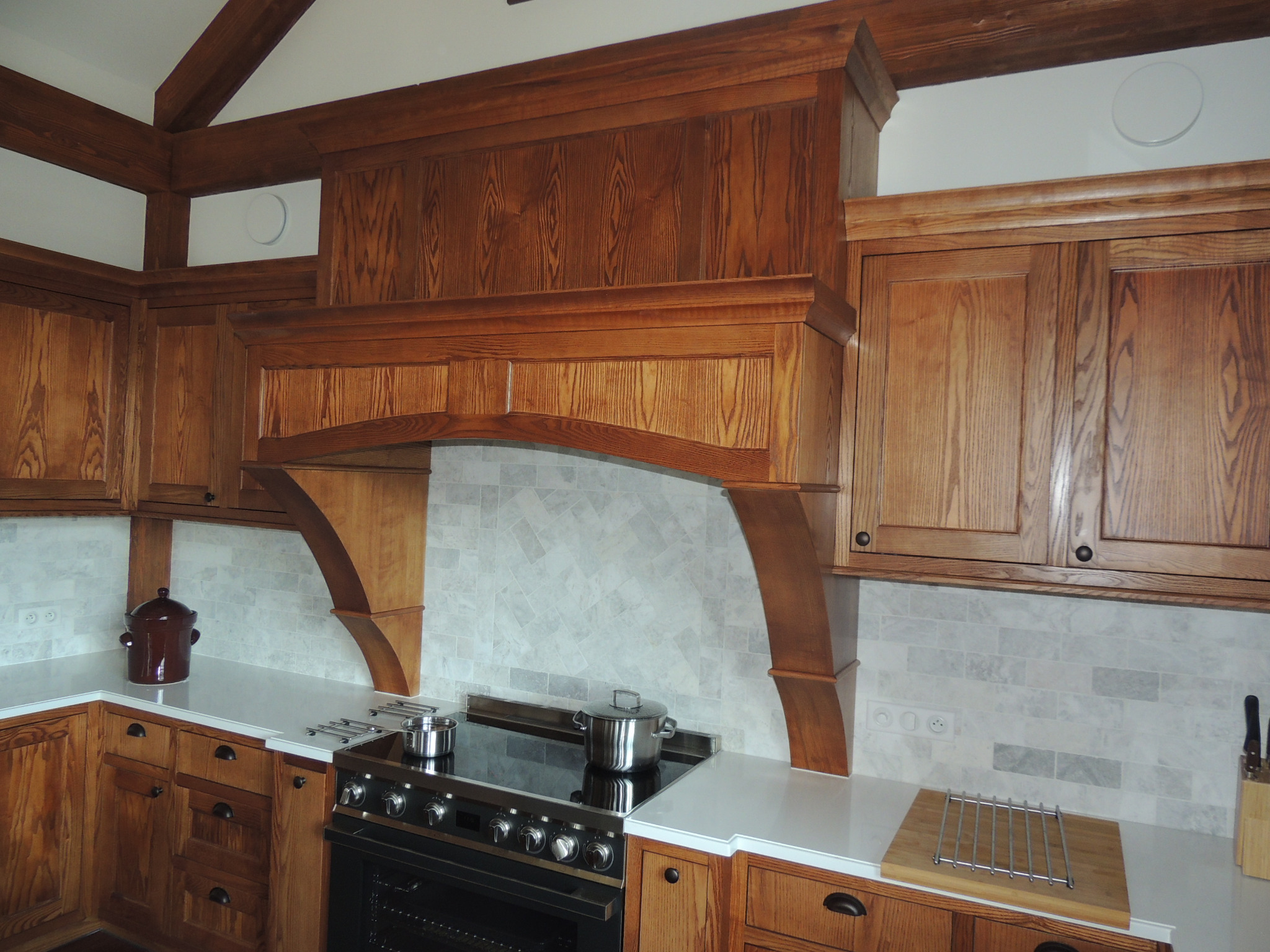 Rustykalna kuchnia z drewnianymi szafkami, okapem i marmurowymi płytkami na ścianie, dwie stalowe patelnie na płycie grzewczej.