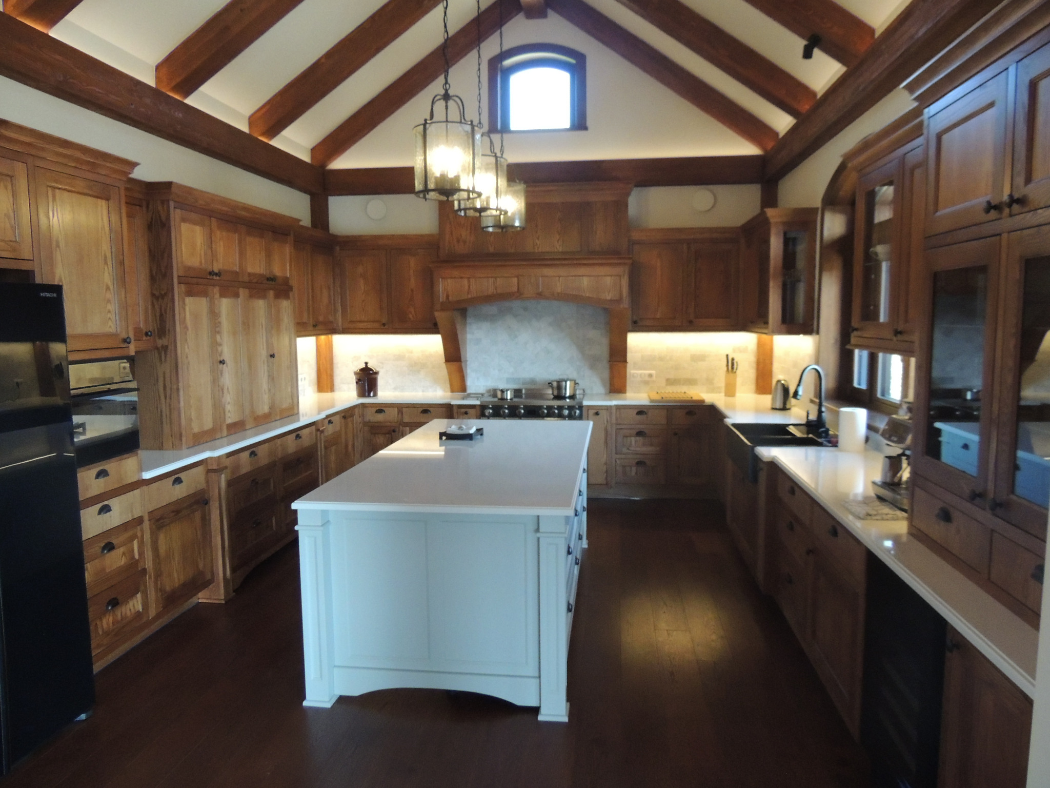 Rustykalna kuchnia z drewnianymi szafkami, białym blatem i wyspą, oświetlona wiszącymi lampami w metalowych obudowach, z drewnianymi belkami stropowymi.