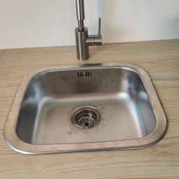 Usługi hydrauliczne - Stalowy zlew kuchenny z zamontowaną baterią, widoczne ślady użytkowania na powierzchni zlewu, blat drewnopodobny i niebieski front szafki pod zlewem.