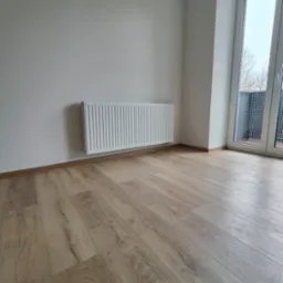 Nowy, biały grzejnik panelowy zamontowany na ścianie w świeżo wykończonym pokoju w Świętochłowicach, z drewnianą podłogą i balkonem widocznym przez okno.