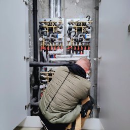 Usługi hydrauliczne - Instalator w trakcie montażu rozdzielacza centralnego ogrzewania w szafce technicznej, widoczne rury, zawory i manometry.