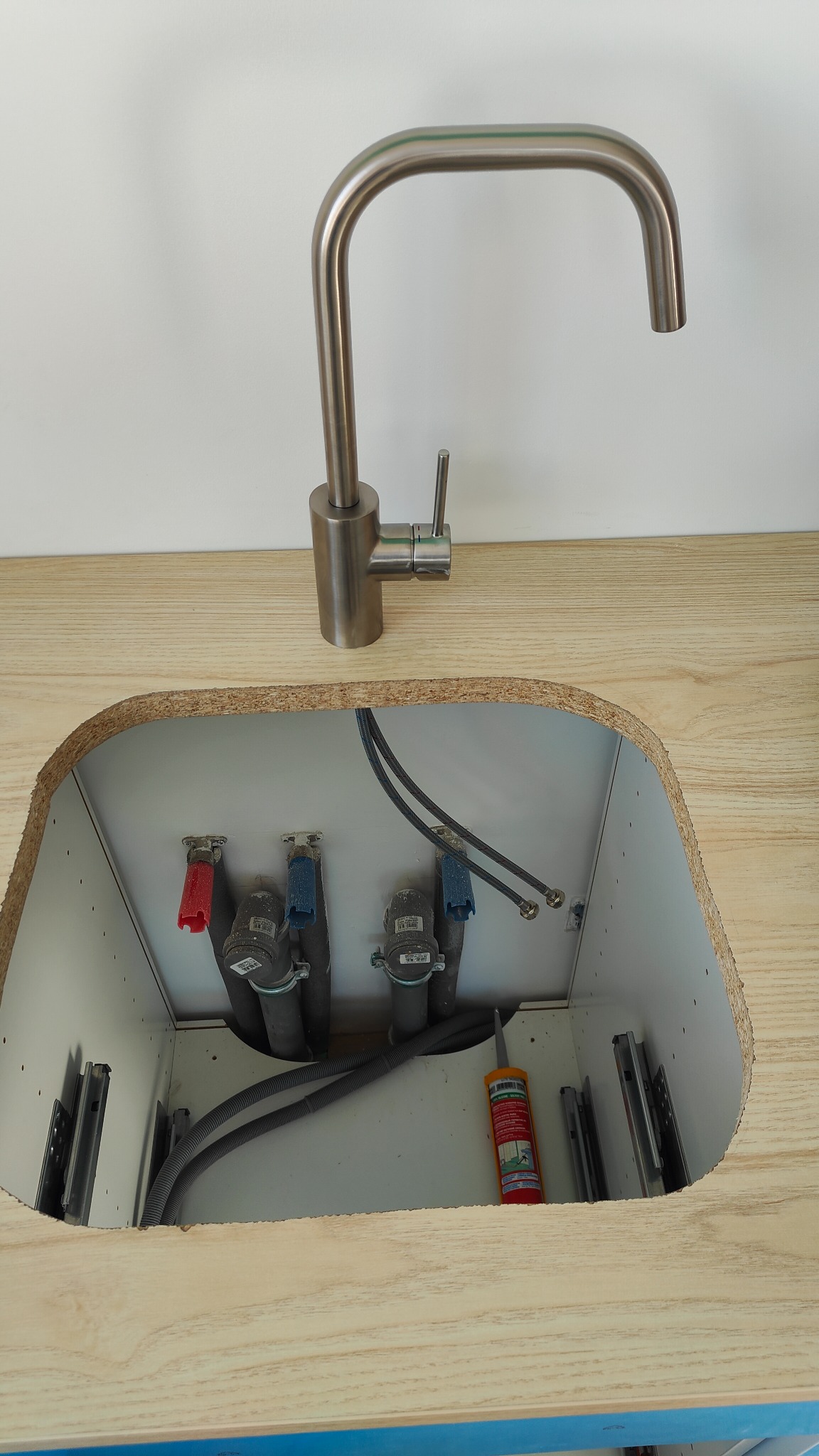 Nowoczesna bateria kuchenna ze stali nierdzewnej zamontowana na blacie z wyciętym otworem, widoczne rury doprowadzające wodę, syfon i tuba silikonu sanitarnego w szafce pod zlewem.