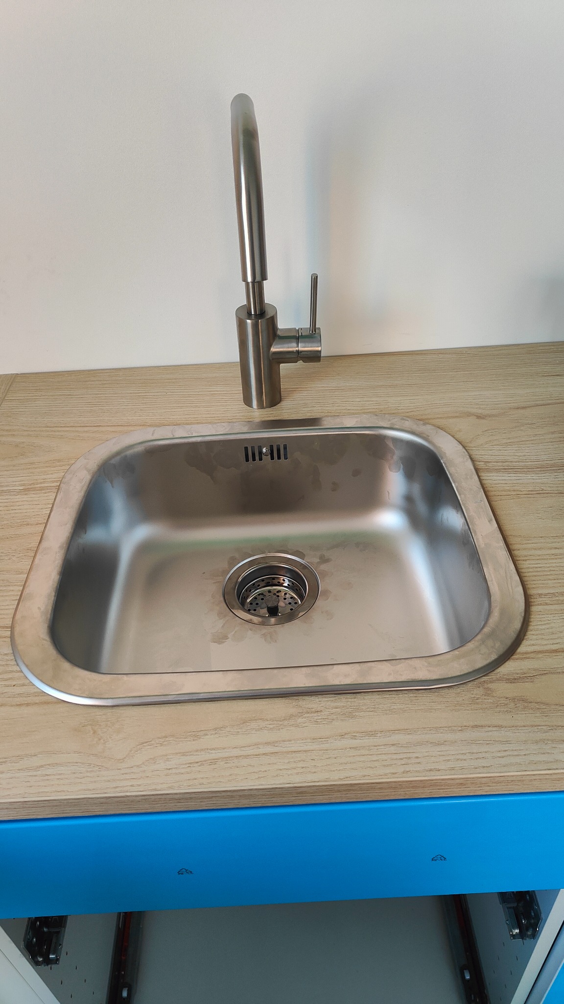 Stalowy zlew kuchenny z zamontowaną baterią, widoczne ślady użytkowania na powierzchni zlewu, blat drewnopodobny i niebieski front szafki pod zlewem.