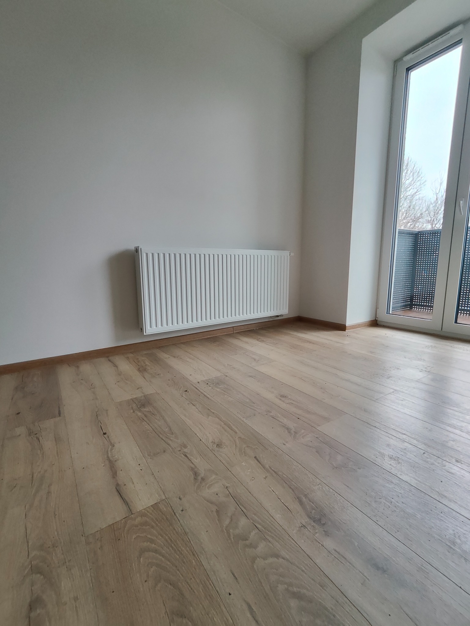 Nowy, biały grzejnik panelowy zamontowany na ścianie w świeżo wykończonym pokoju w Świętochłowicach, z drewnianą podłogą i balkonem widocznym przez okno.