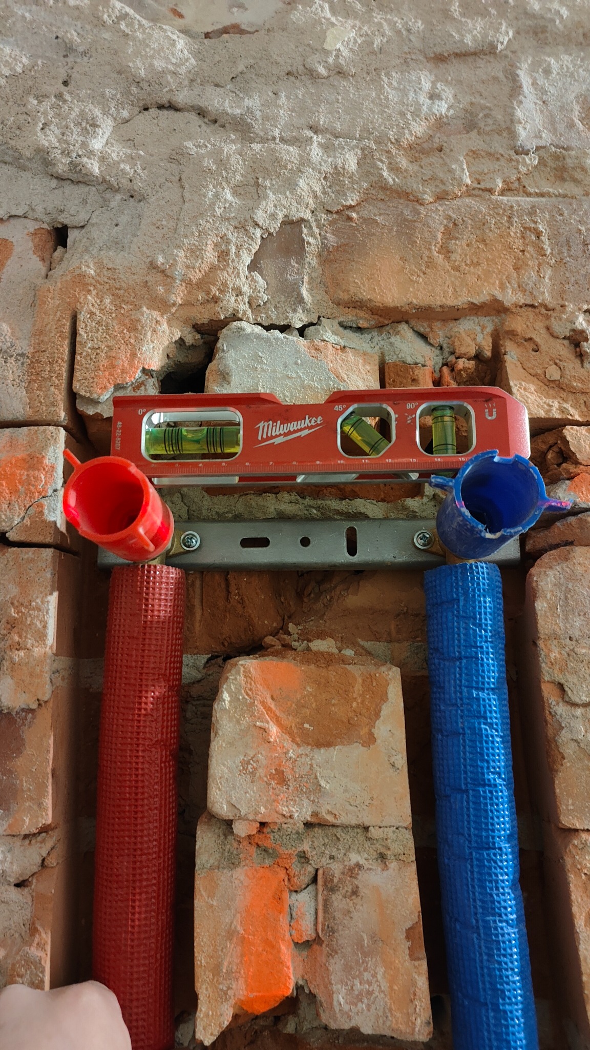 Czerwona i niebieska rura w izolacji zamocowane do ściany z cegły za pomocą metalowej szyny, poziomica Milwaukee na górze, widoczne ślady po kuciu w ścianie.