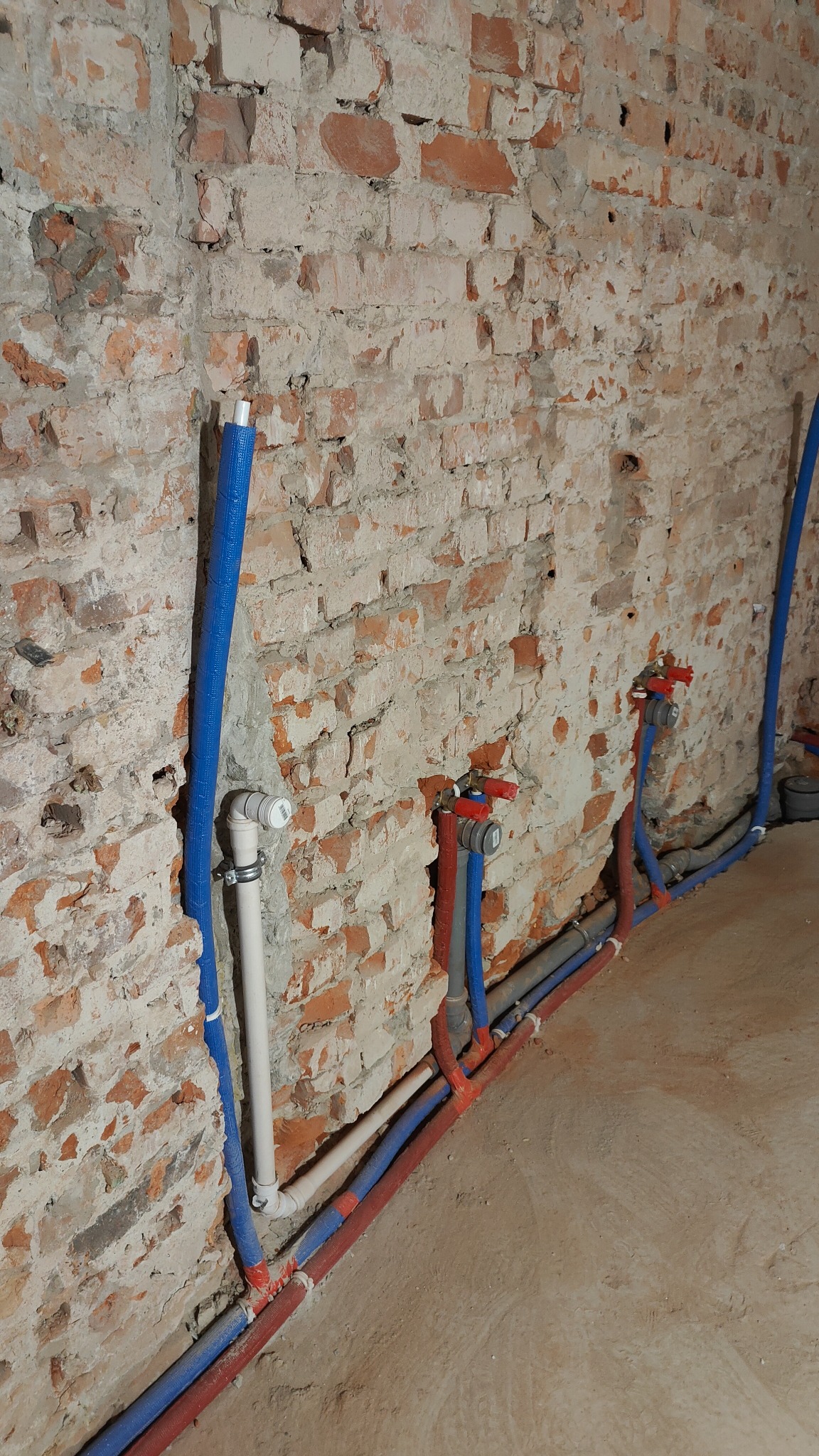 Instalacja wodna z rur PEX w izolacji na tle surowej ściany z cegły, widoczne zawory i złączki.