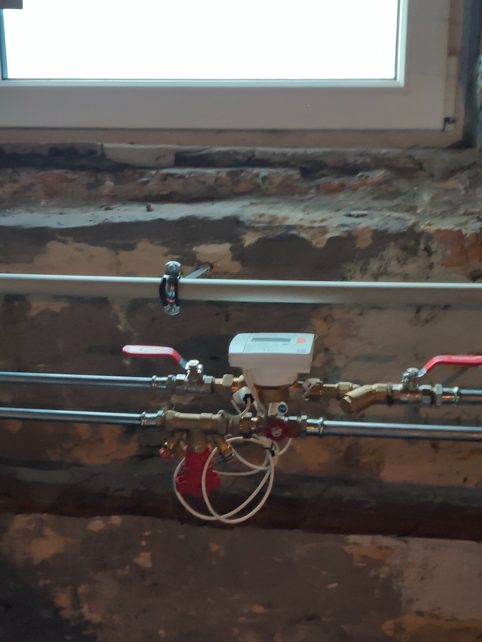 Zamontowany wodomierz z czerwonymi zaworami odcinającymi na tle ściany z oknem.