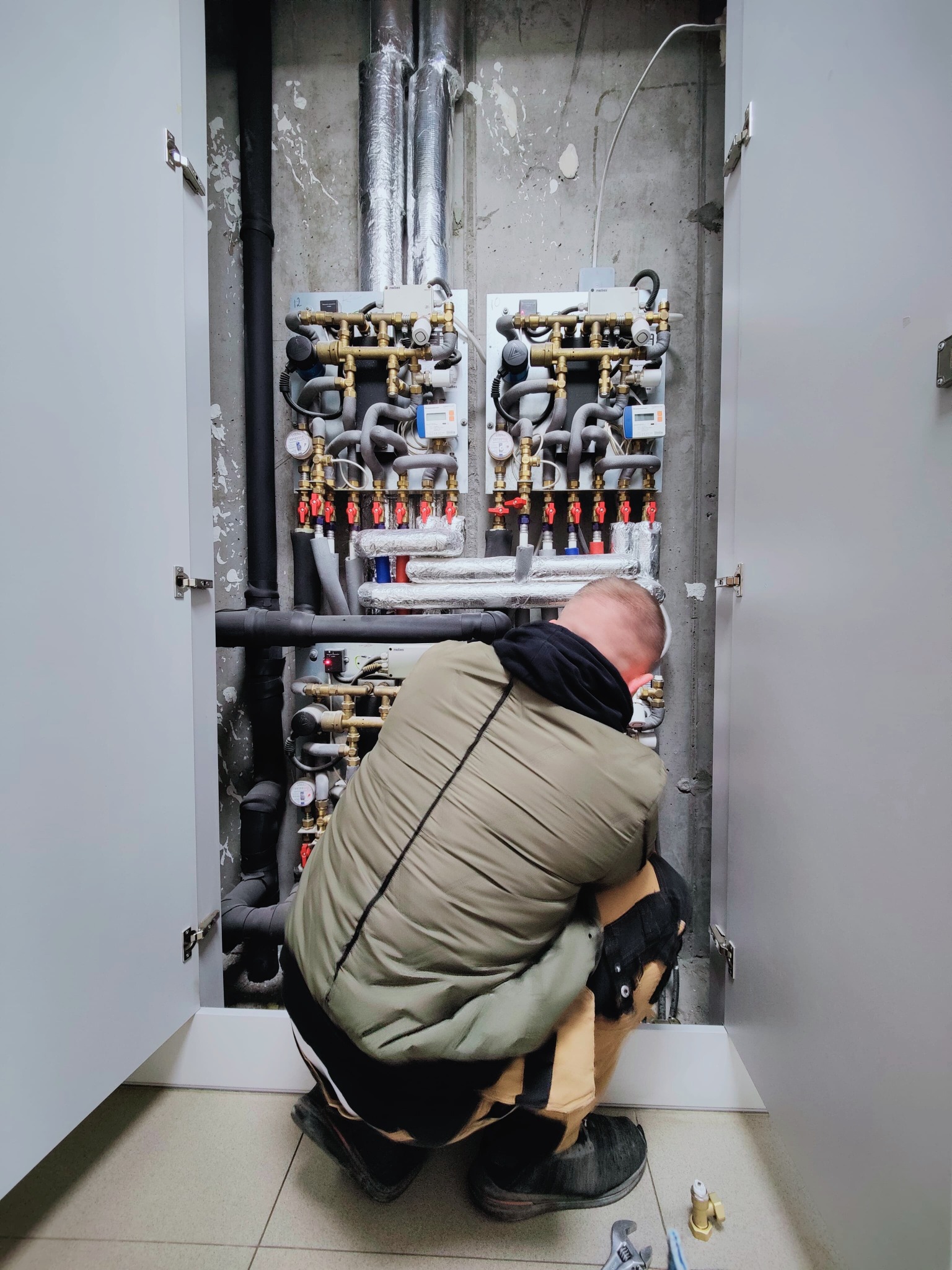 Instalator w trakcie montażu rozdzielacza centralnego ogrzewania w szafce technicznej, widoczne rury, zawory i manometry.