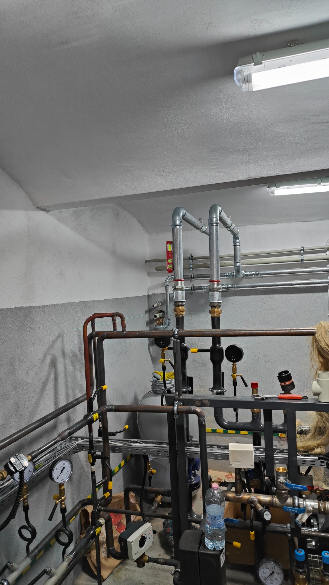 Skomplikowana instalacja rur stalowych i miedzianych w kotłowni, z manometrami, zaworami, butelką wody i poziomnicą, oświetlona lampami jarzeniowymi.