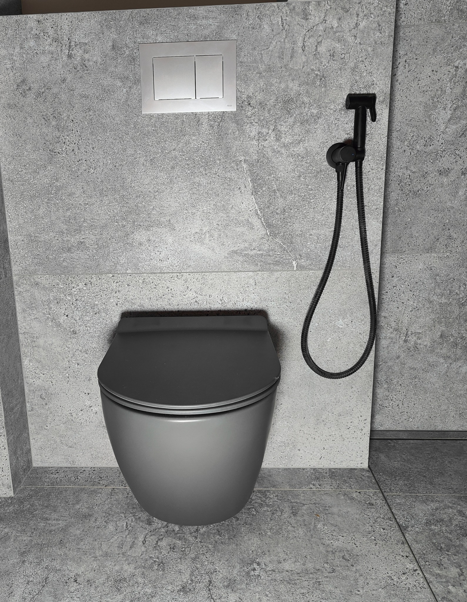 Szara podwieszana toaleta z bidetką i przyciskiem spłukiwania na ścianie wykończonej płytkami imitującymi beton.