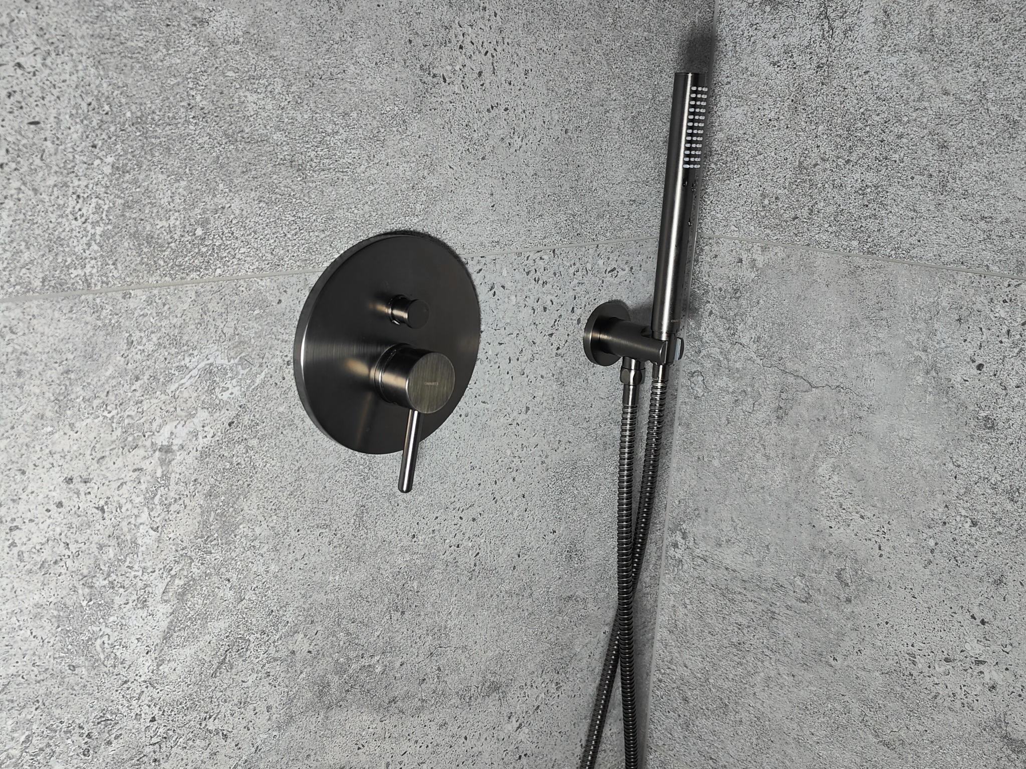 Bateria prysznicowa w kolorze szczotkowanego metalu, zamontowana na szarej, teksturowanej ścianie. Widoczny okrągły panel sterujący i słuchawka prysznicowa z wężem.
