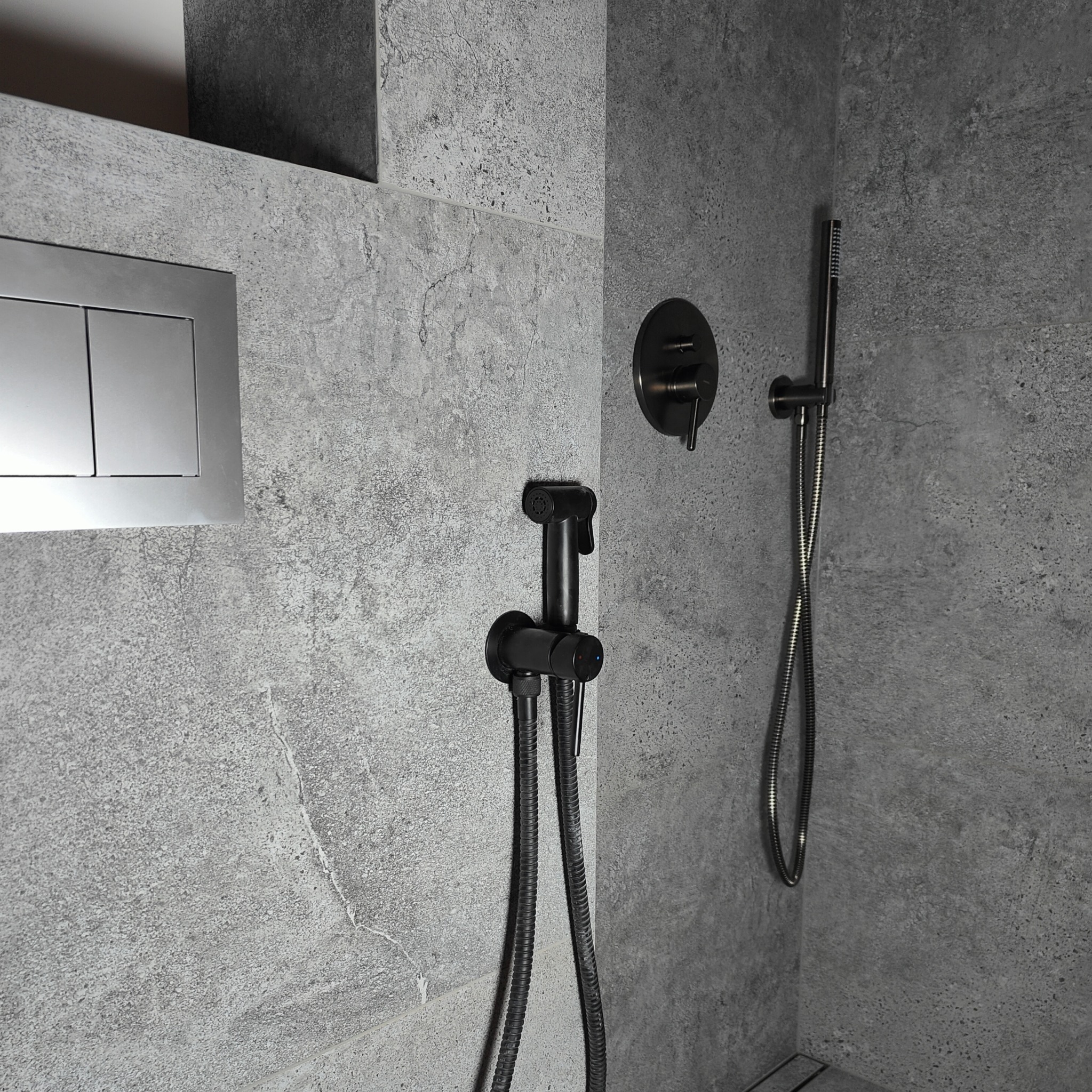 Nowoczesna łazienka z szarą, teksturowaną okładziną, prezentująca podtynkowy panel WC, czarną baterię bidetową z wężem oraz czarną słuchawkę prysznicową z wężem.