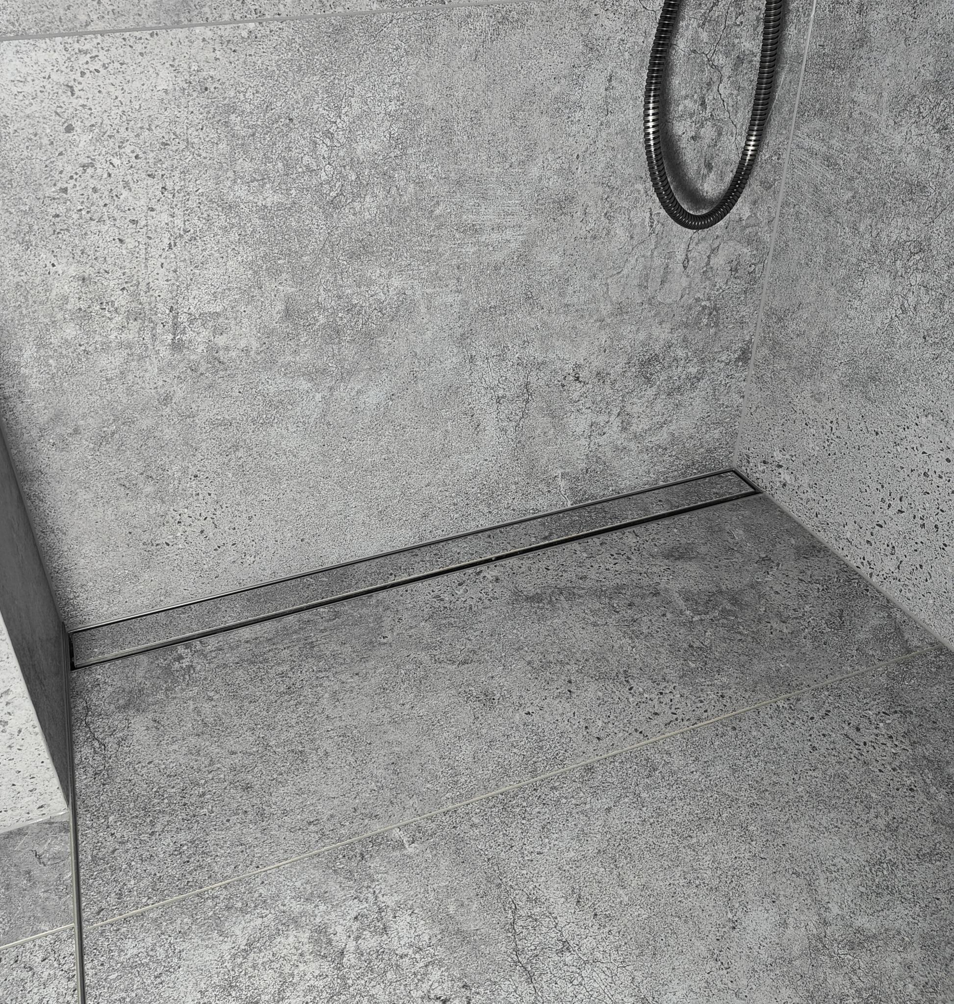Szary odpływ liniowy w posadzce kabiny prysznicowej z szarej, chropowatej płyty. Widoczny wąż prysznicowy.