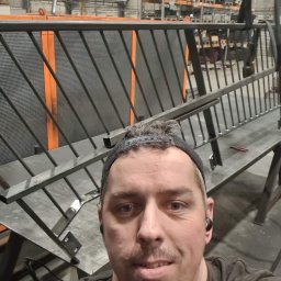 Bartosz Zając - Selfie pracownika na tle hali produkcyjnej z częściowo zmontowaną metalową konstrukcją z ukośnymi prętami i siatką, prawdopodobnie elementu ogrodzenia lub balustrady, widoczny suwnica w tle.