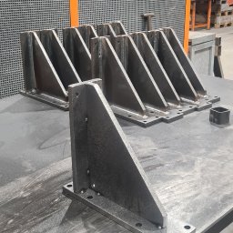 Bartosz Zając - Surowe, spawane stalowe wsporniki kątowe z otworami na śruby, ułożone na metalowym stole w warsztacie, widoczne ślady spawania i obróbki.