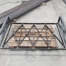 Bartosz Zając - Surowa, metalowa konstrukcja balustrady z geometrycznym wzorem rombów, umieszczona na drewnianej palecie w hali produkcyjnej.