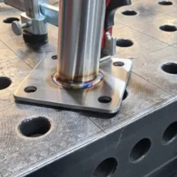 Detal spoiny rury ze stalową płytką, widoczne ślady spawania TIG, mocowane zaciskiem na perforowanym stole spawalniczym.