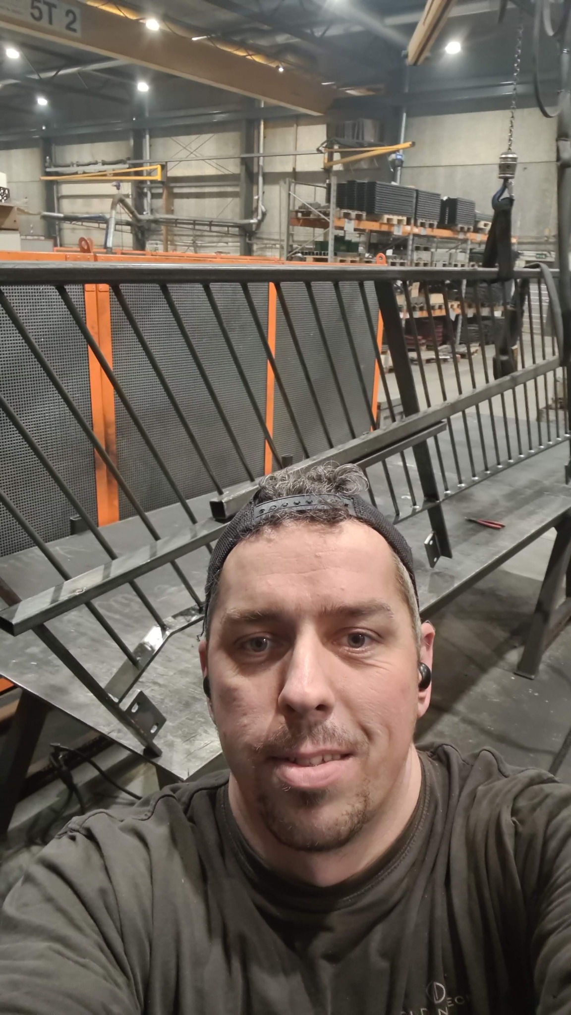Selfie pracownika na tle hali produkcyjnej z częściowo zmontowaną metalową konstrukcją z ukośnymi prętami i siatką, prawdopodobnie elementu ogrodzenia lub balustrady, widoczny suwnica w tle.
