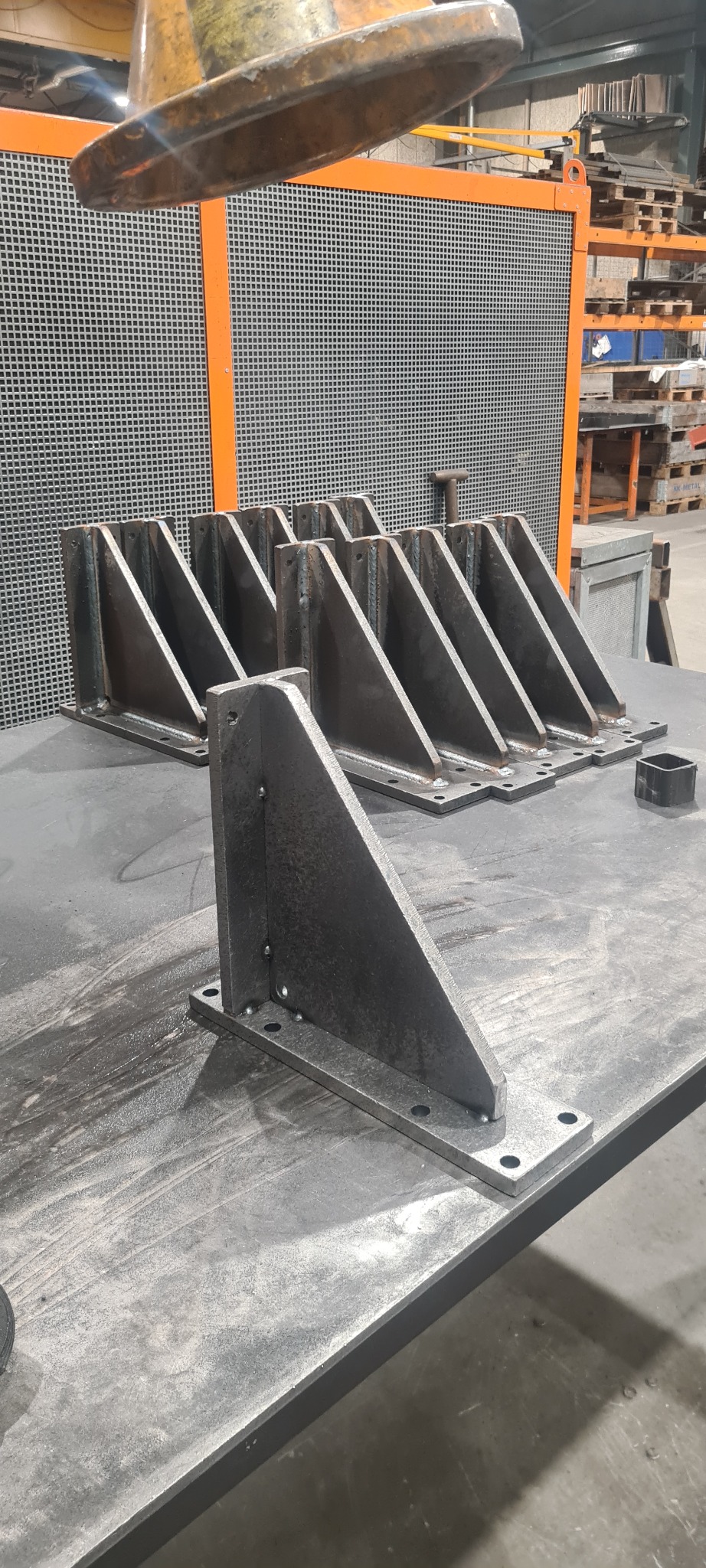 Surowe, spawane stalowe wsporniki kątowe z otworami na śruby, ułożone na metalowym stole w warsztacie, widoczne ślady spawania i obróbki.