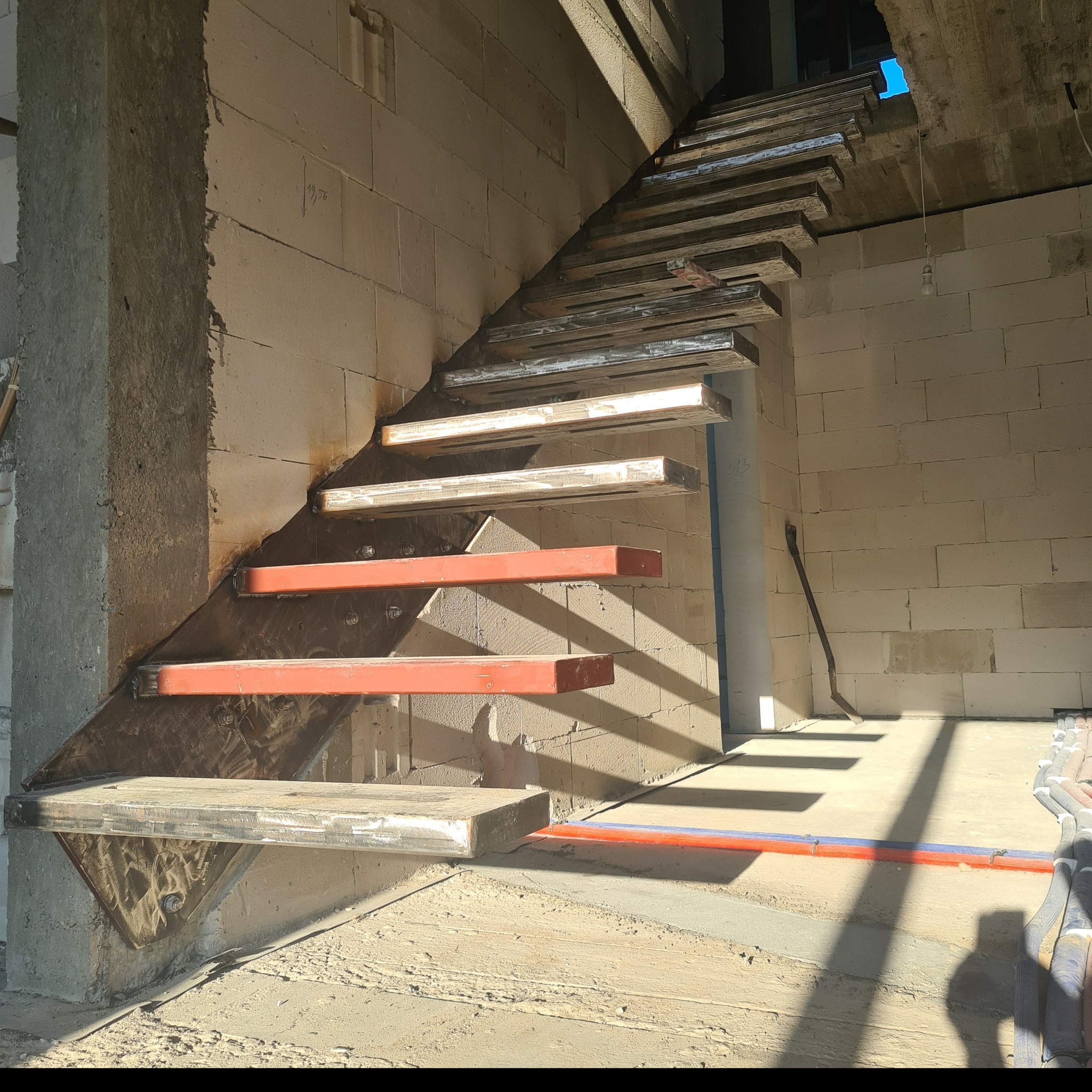 Surowe, metalowe schody z czerwonymi elementami konstrukcyjnymi, zamontowane w surowym wnętrzu budynku w trakcie budowy, z widocznymi blokami betonowymi i przewodami instalacyjnymi.