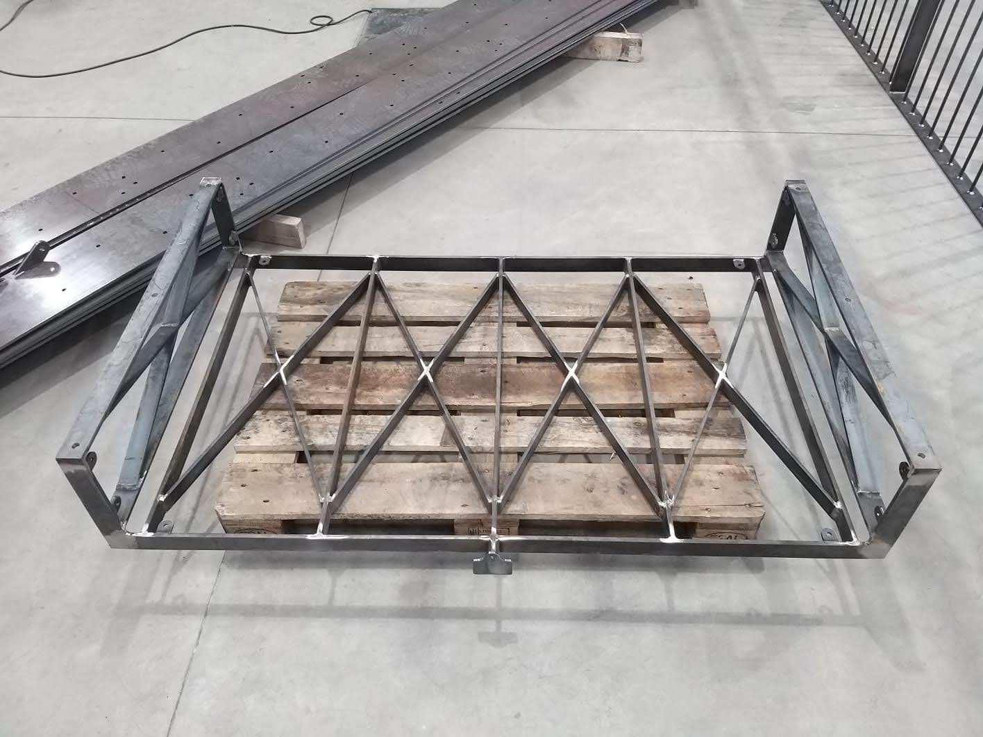 Surowa, metalowa konstrukcja balustrady z geometrycznym wzorem rombów, umieszczona na drewnianej palecie w hali produkcyjnej.