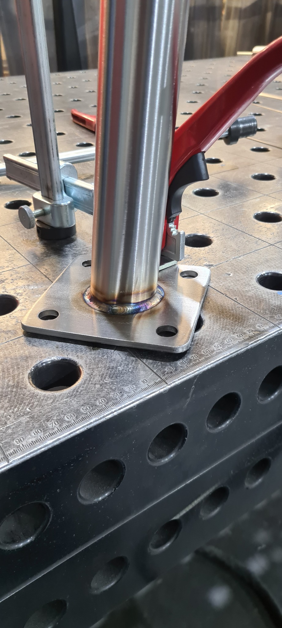 Detal spoiny rury ze stalową płytką, widoczne ślady spawania TIG, mocowane zaciskiem na perforowanym stole spawalniczym.