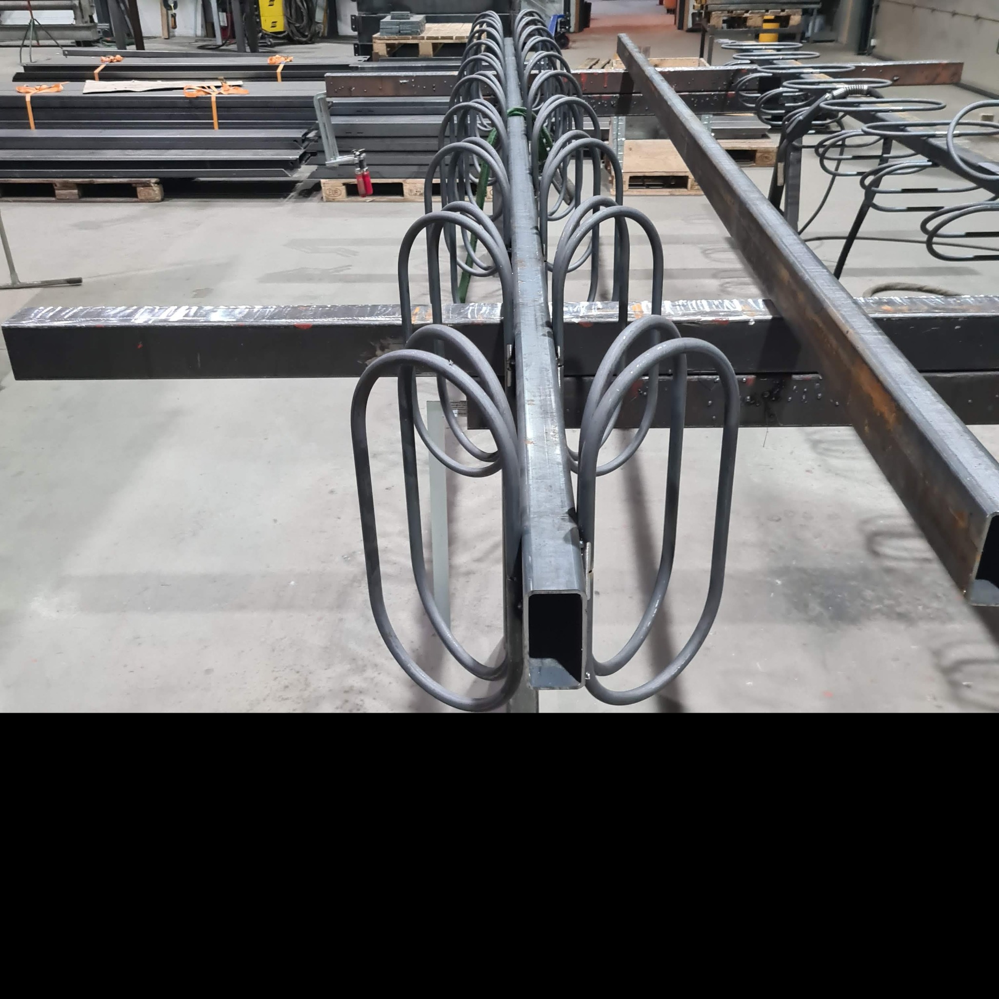 Surowy metalowy stelaż z prętami w kształcie pętli, prawdopodobnie element konstrukcyjny, widoczny w hali produkcyjnej z innymi metalowymi profilami w tle.