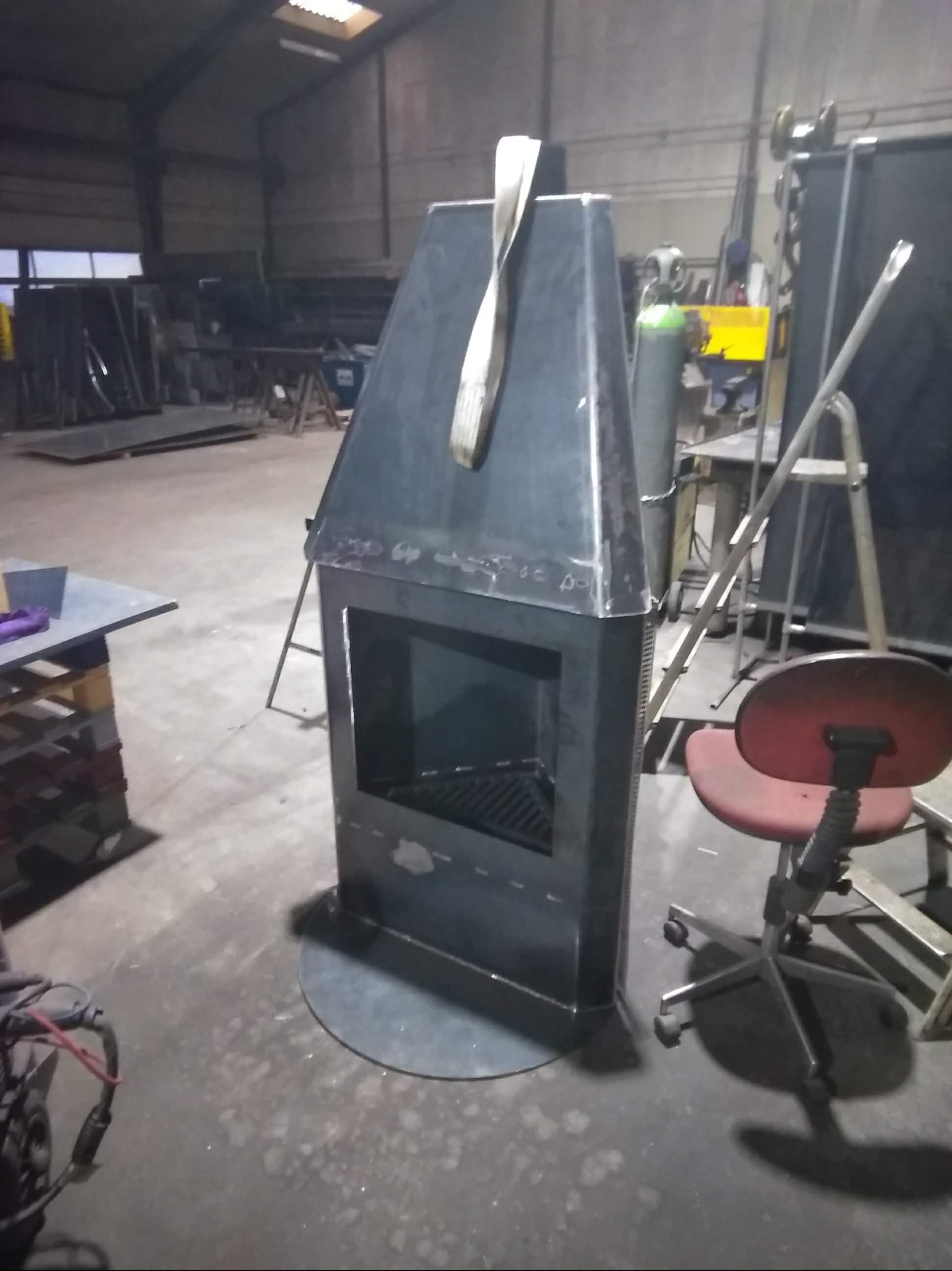 Surowy, metalowy kominek zewnętrzny w trakcie produkcji w warsztacie, z widocznymi śladami spawania i elementami wyposażenia warsztatowego w tle.