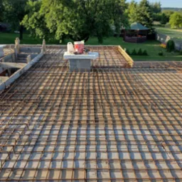 Zbrojenie płyty fundamentowej domu jednorodzinnego z widocznymi deskami szalunkowymi, na tle zielonego ogrodu z altaną.