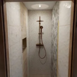 Nowoczesna kabina prysznicowa z armaturą w kolorze miedzi, wykończona płytkami w odcieniach beżu, bieli i szarości z subtelnym wzorem, oświetlona punktowym światłem.