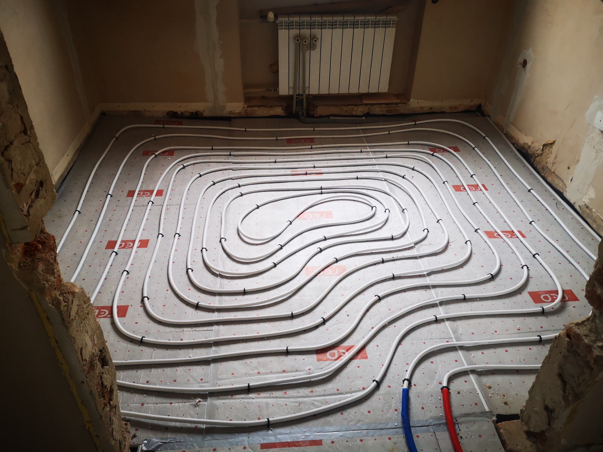 Rozłożona instalacja ogrzewania podłogowego z białych rurek na izolacji, widoczne fragmenty ściany w stanie surowym i grzejnik na ścianie.