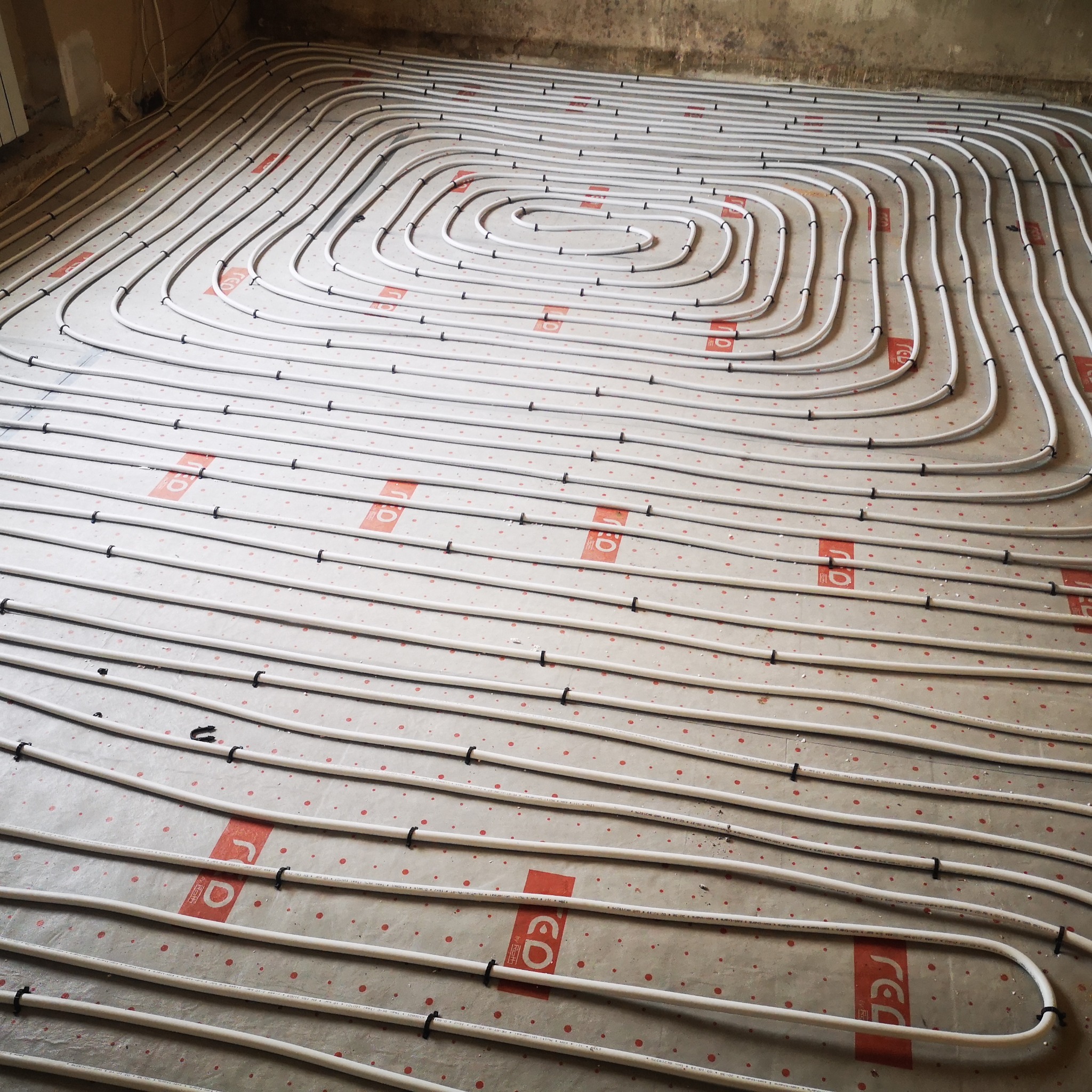 Układ ogrzewania podłogowego z białych rur, zamocowanych czarnymi uchwytami na folii z czerwonym nadrukiem, widok z góry na surową podłogę.