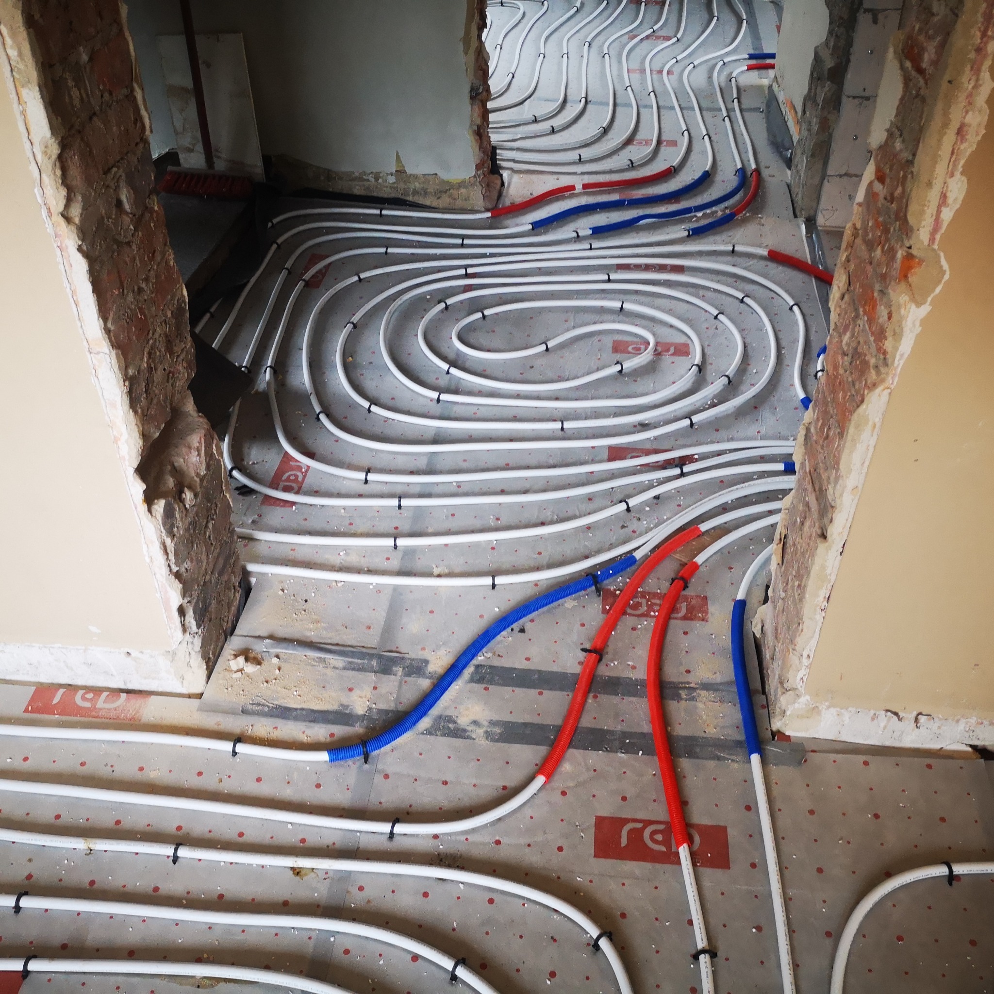 Instalacja ogrzewania podłogowego w trakcie remontu w Psarach, widoczne białe rury grzewcze ułożone spiralnie na podkładzie, z czerwonymi i niebieskimi rurami doprowadzającymi, fragmenty ścian...