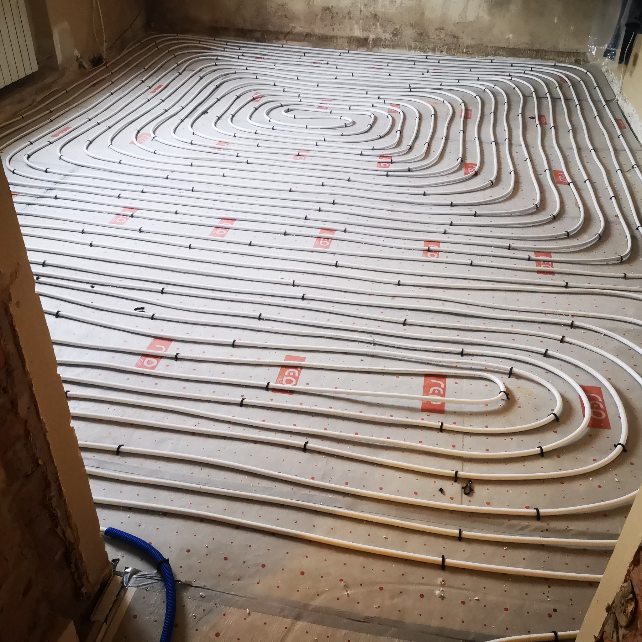 Rozłożone rury ogrzewania podłogowego na folii izolacyjnej z czerwonymi oznaczeniami, przymocowane czarnymi uchwytami do podłoża, widoczne fragmenty ścian w surowym stanie.