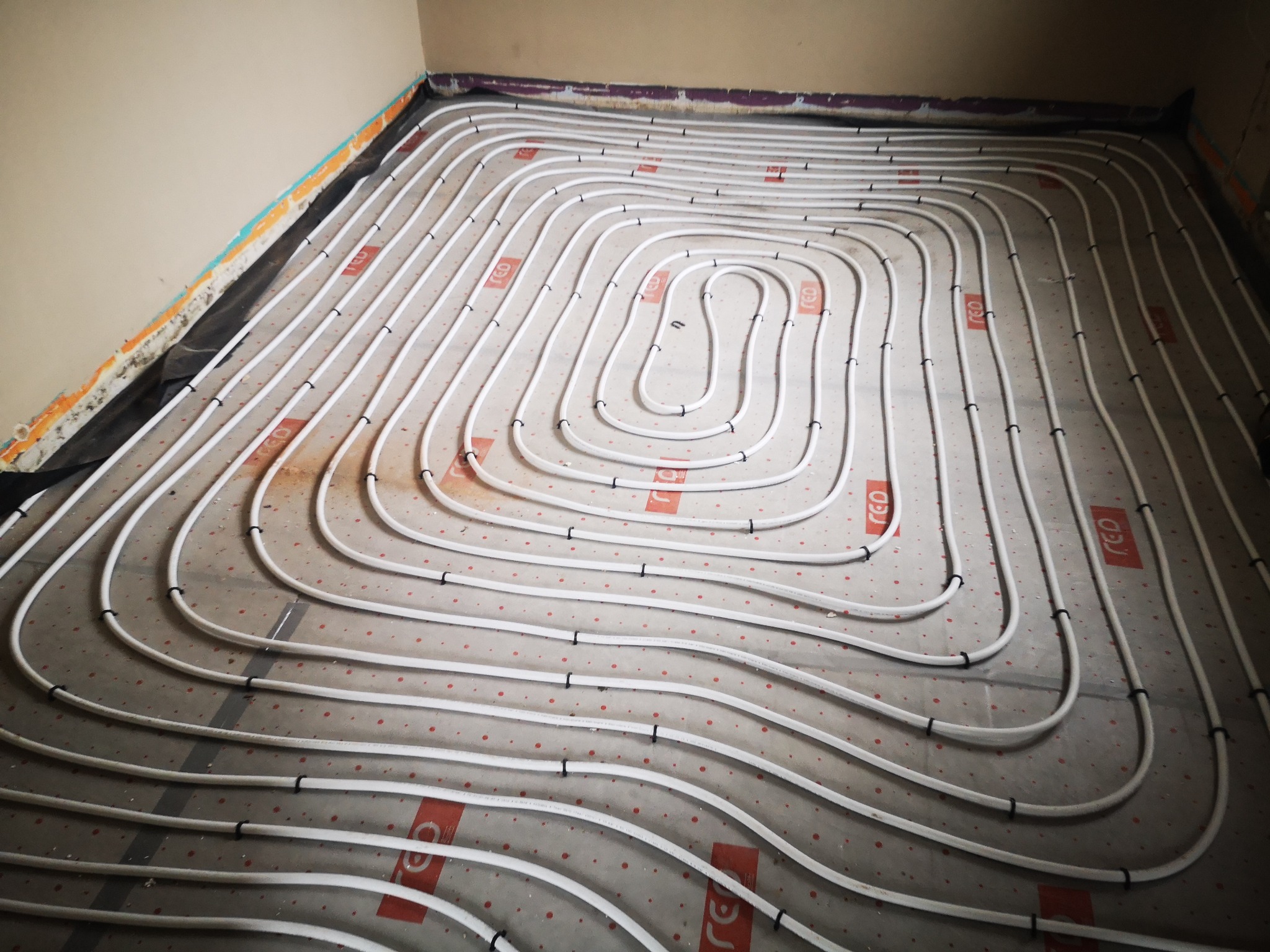Rozłożona instalacja ogrzewania podłogowego z białych rur mocowanych czarnymi opaskami na płytach izolacyjnych z czerwonym nadrukiem w pomieszczeniu w trakcie prac wykończeniowych.