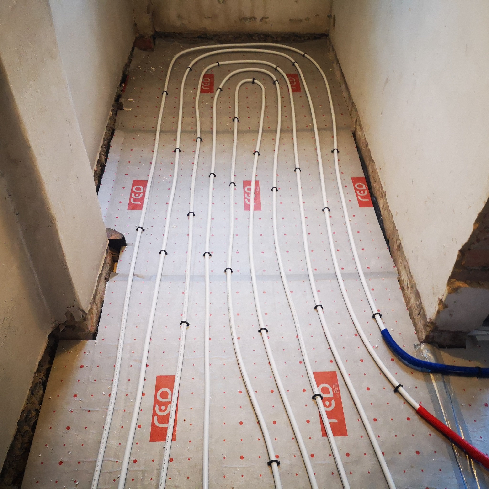 Białe rury ogrzewania podłogowego ułożone w pętle na izolacji z czerwonym nadrukiem, widoczne fragmenty ścian w stanie surowym.