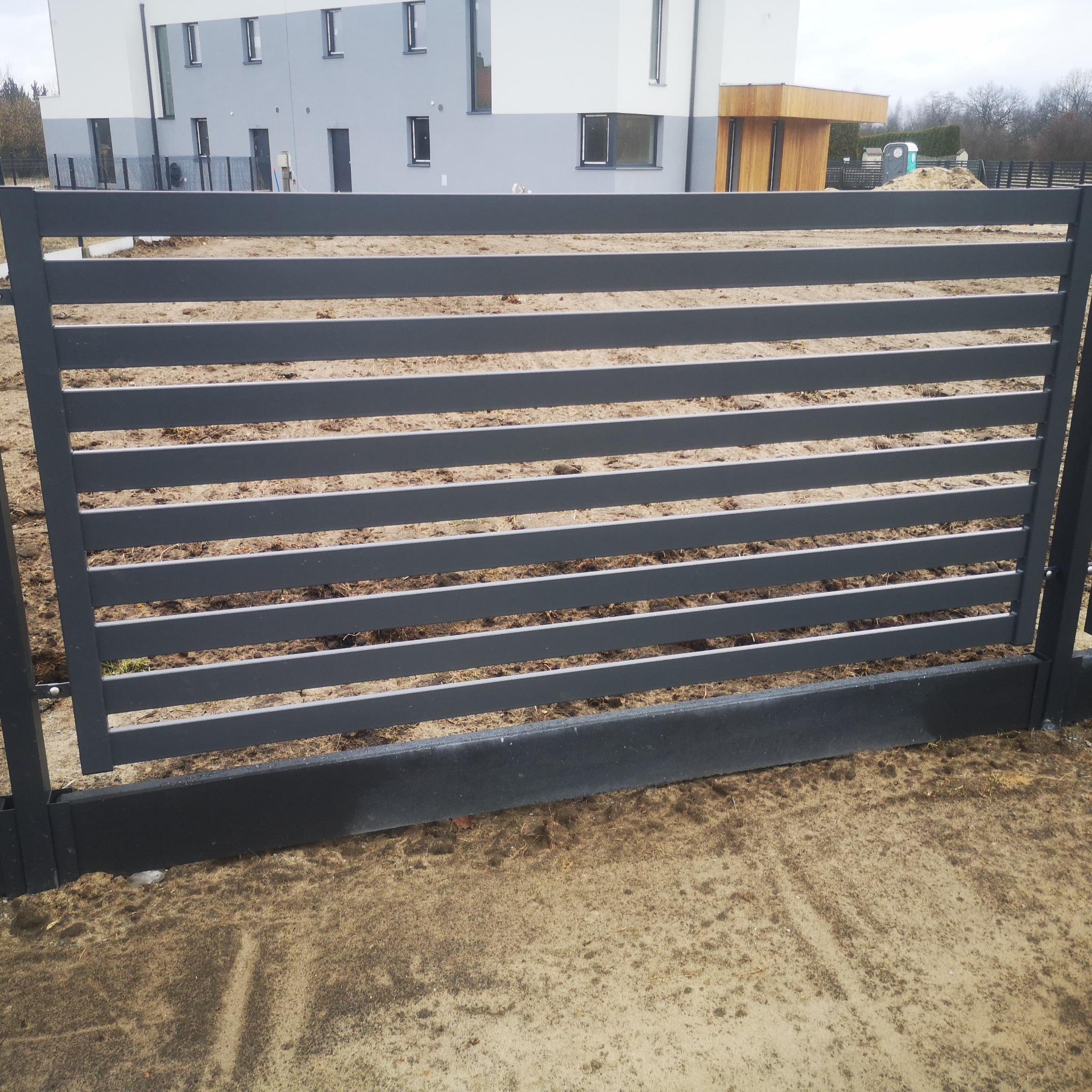 Nowoczesna, pozioma, szara brama wjazdowa na posesję, z widocznym w tle budynkiem w trakcie budowy i przenośną toaletą.