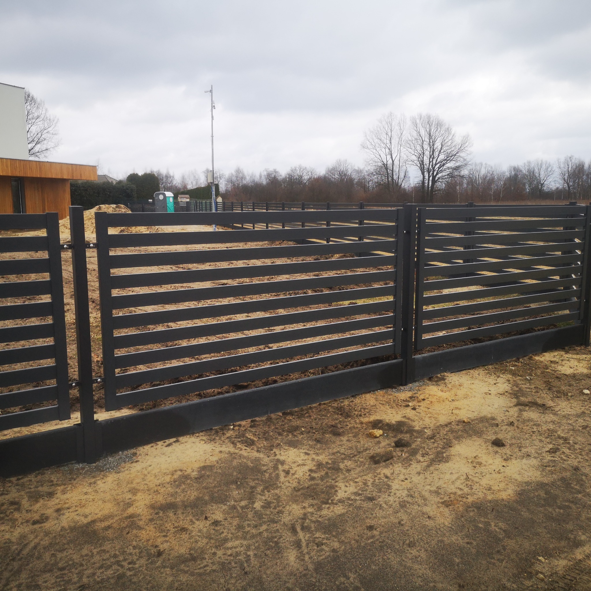 Nowoczesne, poziome ogrodzenie panelowe w kolorze grafitowym, z widocznym fragmentem budynku z drewnianą elewacją i tymczasową toaletą na posesji, na tle pochmurnego nieba.