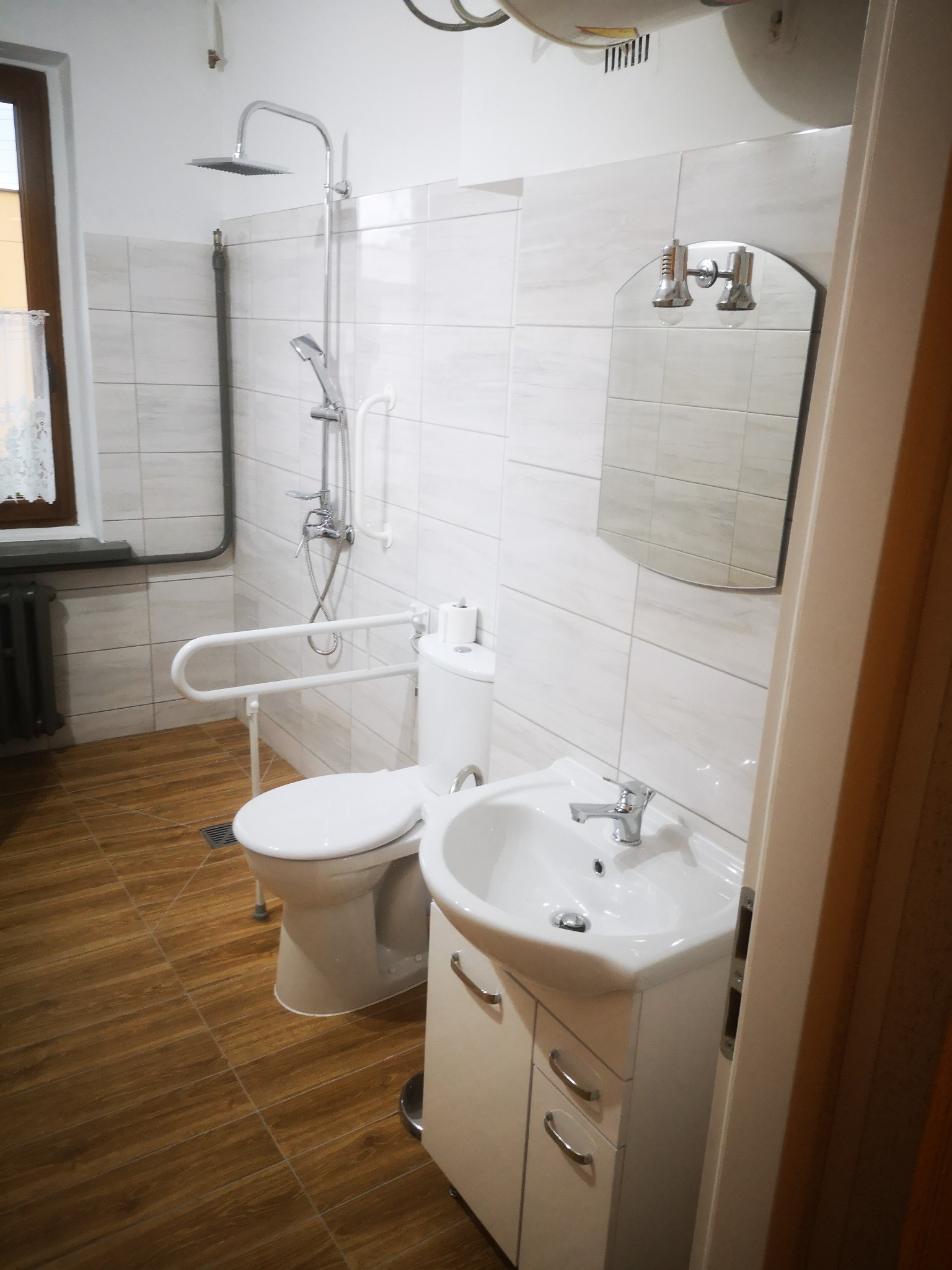 Wykończona łazienka z białymi kafelkami, brązowymi płytkami podłogowymi imitującymi drewno, umywalką z szafką, toaletą z uchwytem dla osób niepełnosprawnych oraz zestawem prysznicowym z deszczownicą.