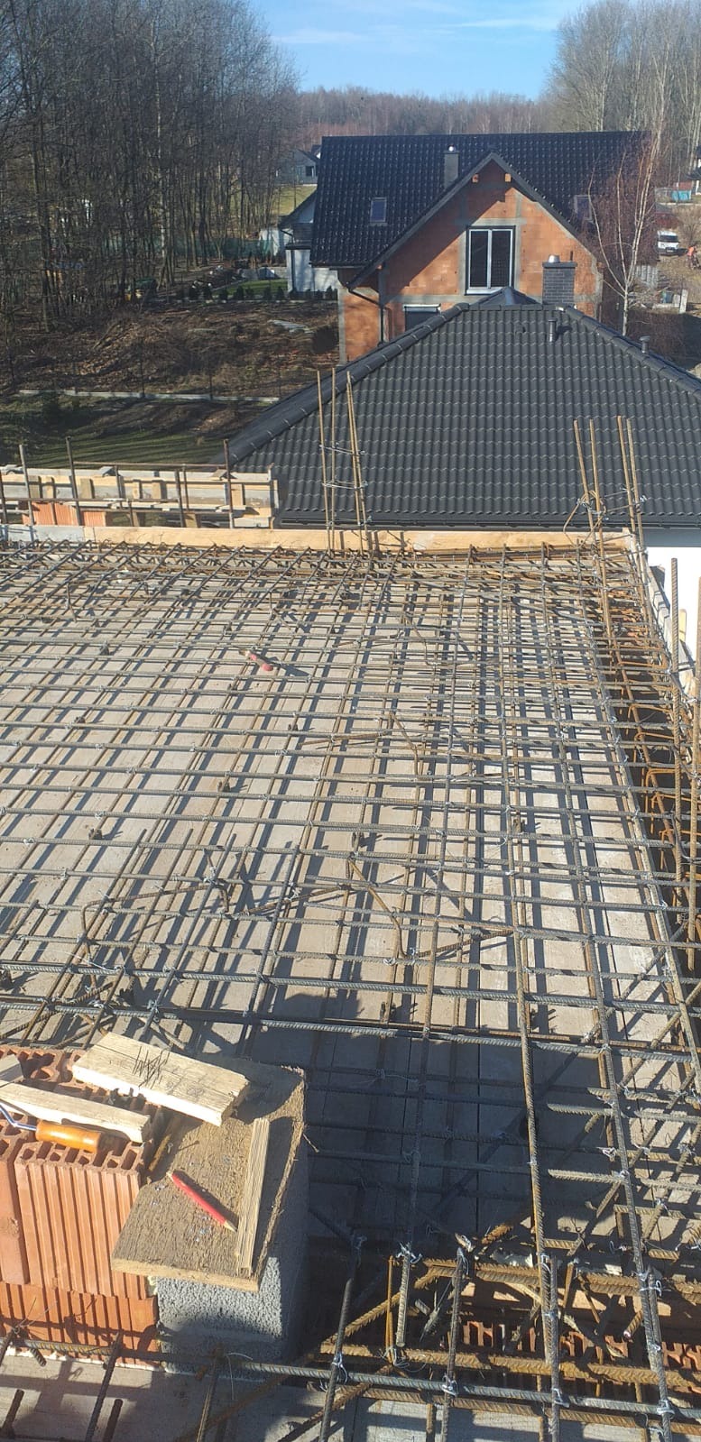Zbrojenie płyty fundamentowej domu jednorodzinnego, widok z góry na siatkę z prętów stalowych, w tle budynek w stanie surowym otwartym i drzewa.