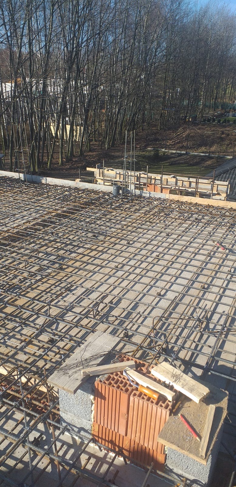 Zbrojenie płyty fundamentowej z widocznymi elementami szalunku i cegłami na obrzeżach, w tle drzewa bez liści.