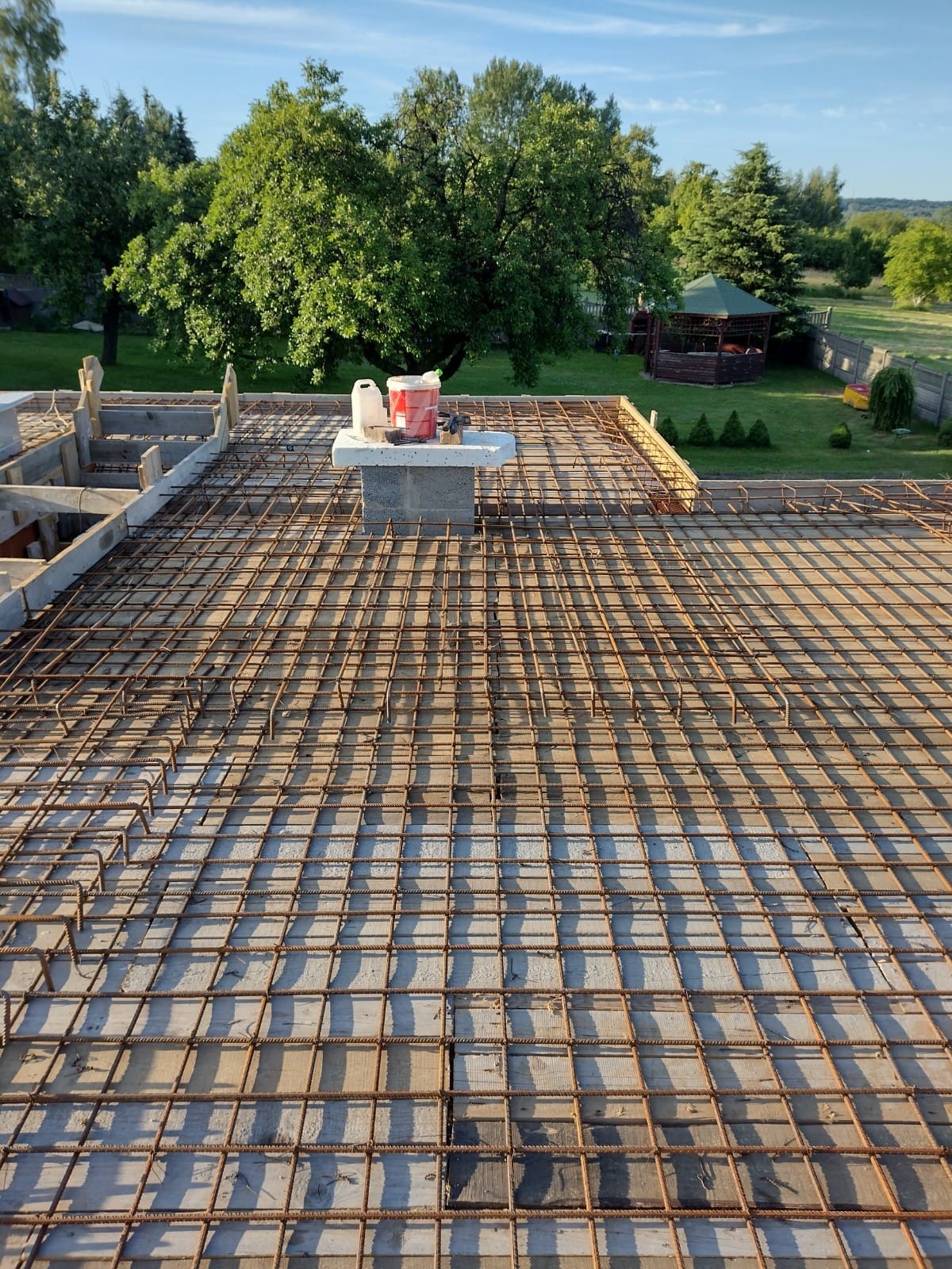 Zbrojenie płyty fundamentowej domu jednorodzinnego z widocznymi deskami szalunkowymi, na tle zielonego ogrodu z altaną.