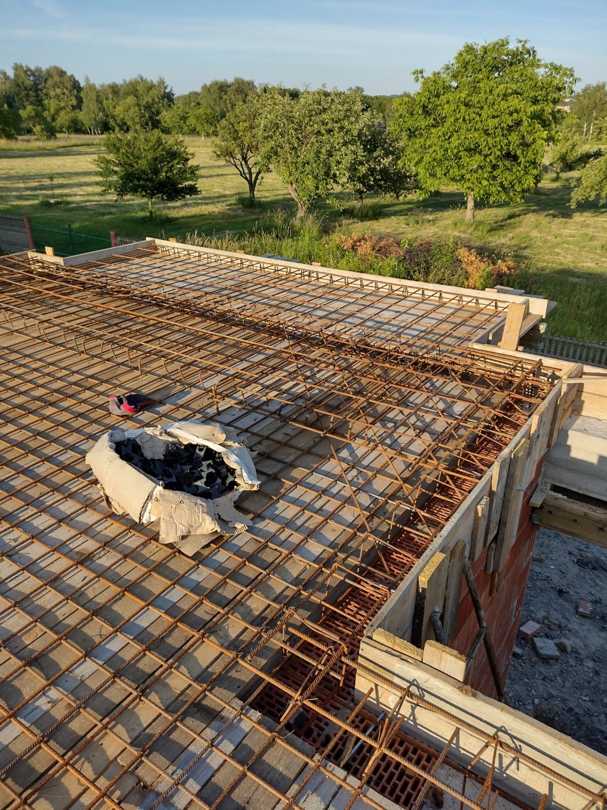 Zbrojenie stropu domu jednorodzinnego z widoczną siatką z prętów stalowych, szalunkiem i kartonem ze spinkami montażowymi, w tle zieleń drzew.