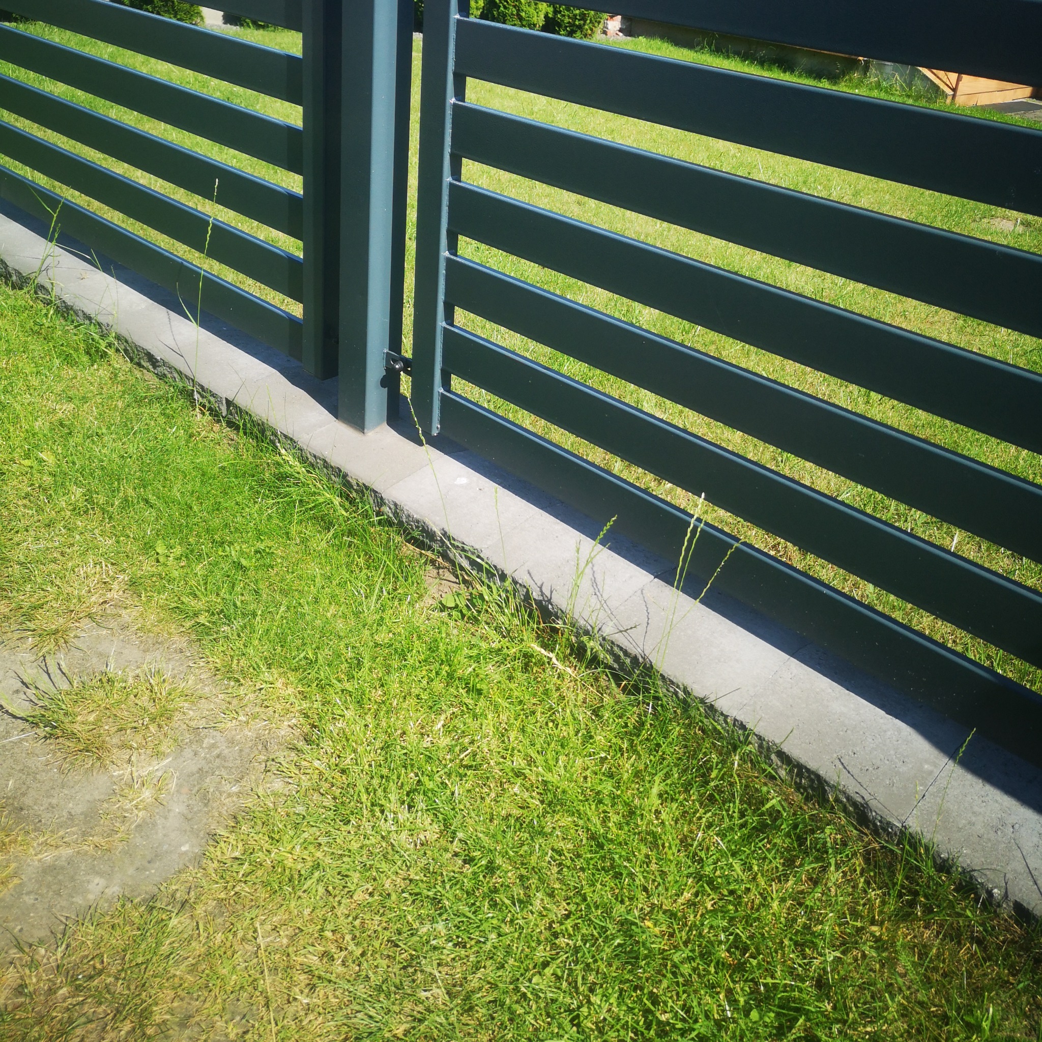 Nowoczesne, poziome przęsła ogrodzenia panelowego w kolorze grafitowym, osadzone na betonowej podmurówce, z widoczną zieloną trawą.