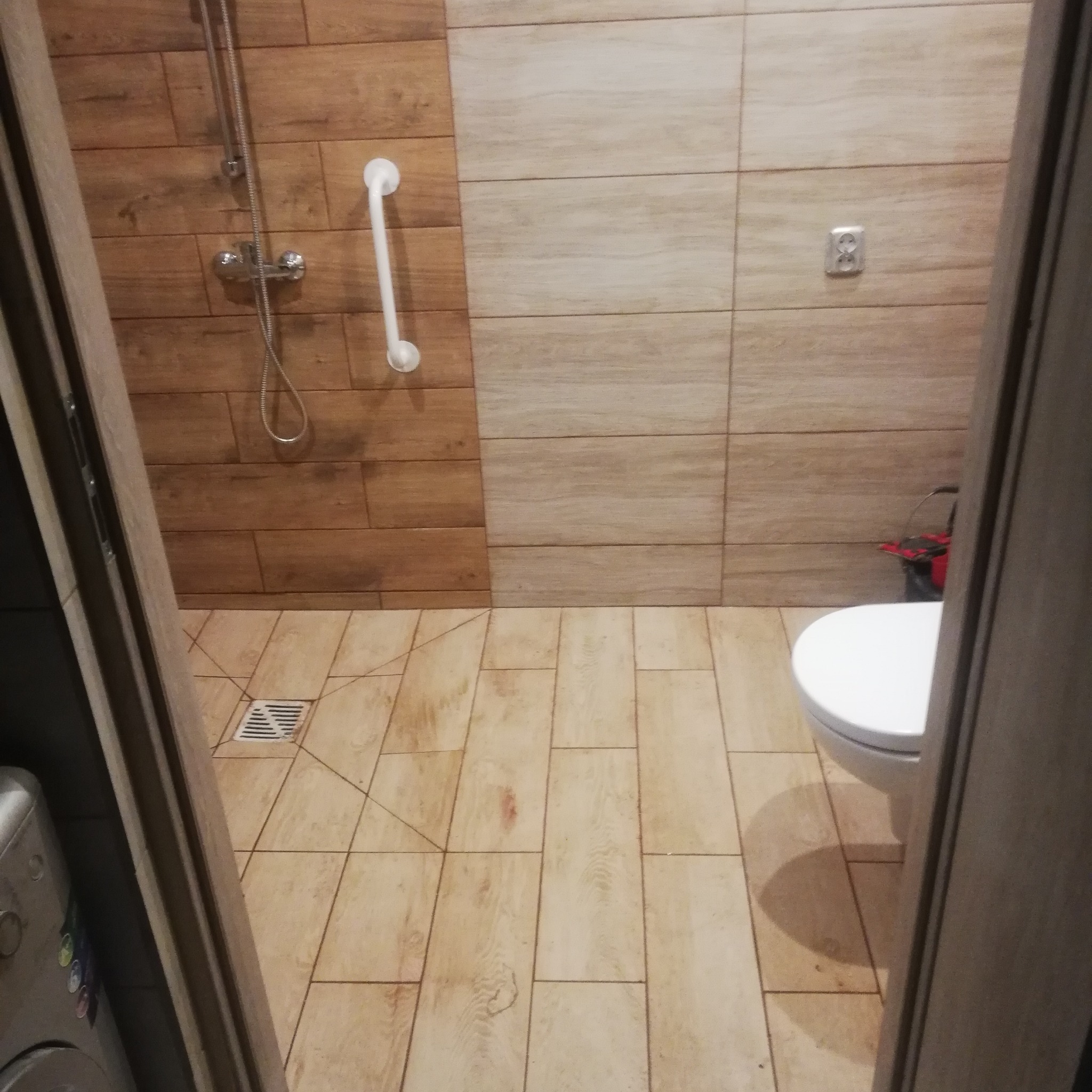 Wykończona łazienka z brązowymi i beżowymi płytkami imitującymi drewno, z zamontowanym uchwytem dla niepełnosprawnych przy prysznicu i widocznym odpływem liniowym w podłodze.