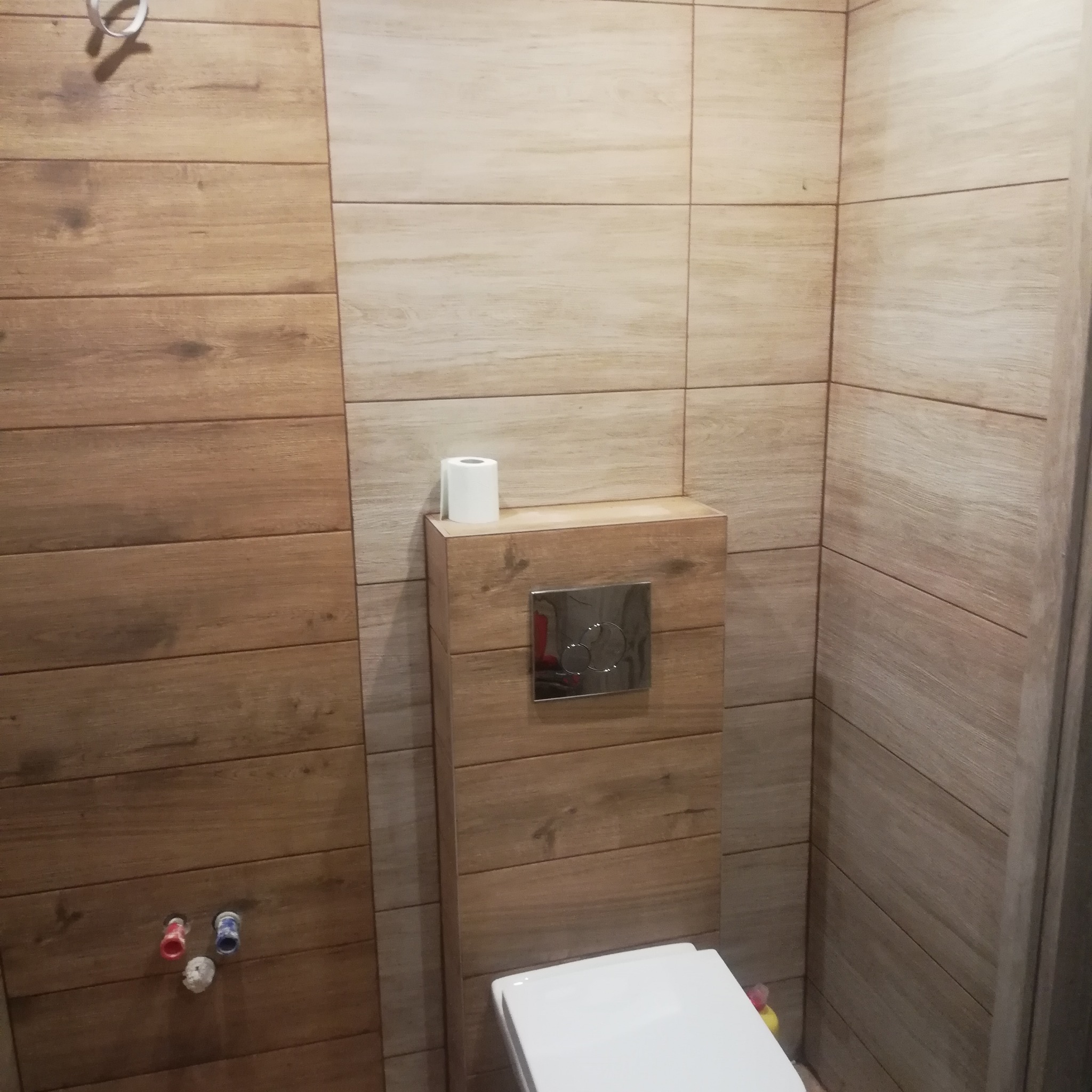 Nowoczesna łazienka z toaletą podwieszaną, obudowaną drewnopodobnymi płytkami, z rolką papieru toaletowego na obudowie.