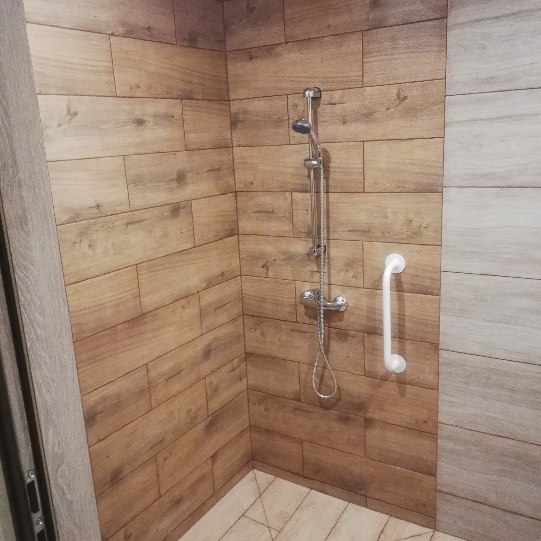 Kabina prysznicowa wyłożona płytkami imitującymi drewno, z zamontowaną baterią i uchwytem dla niepełnosprawnych.