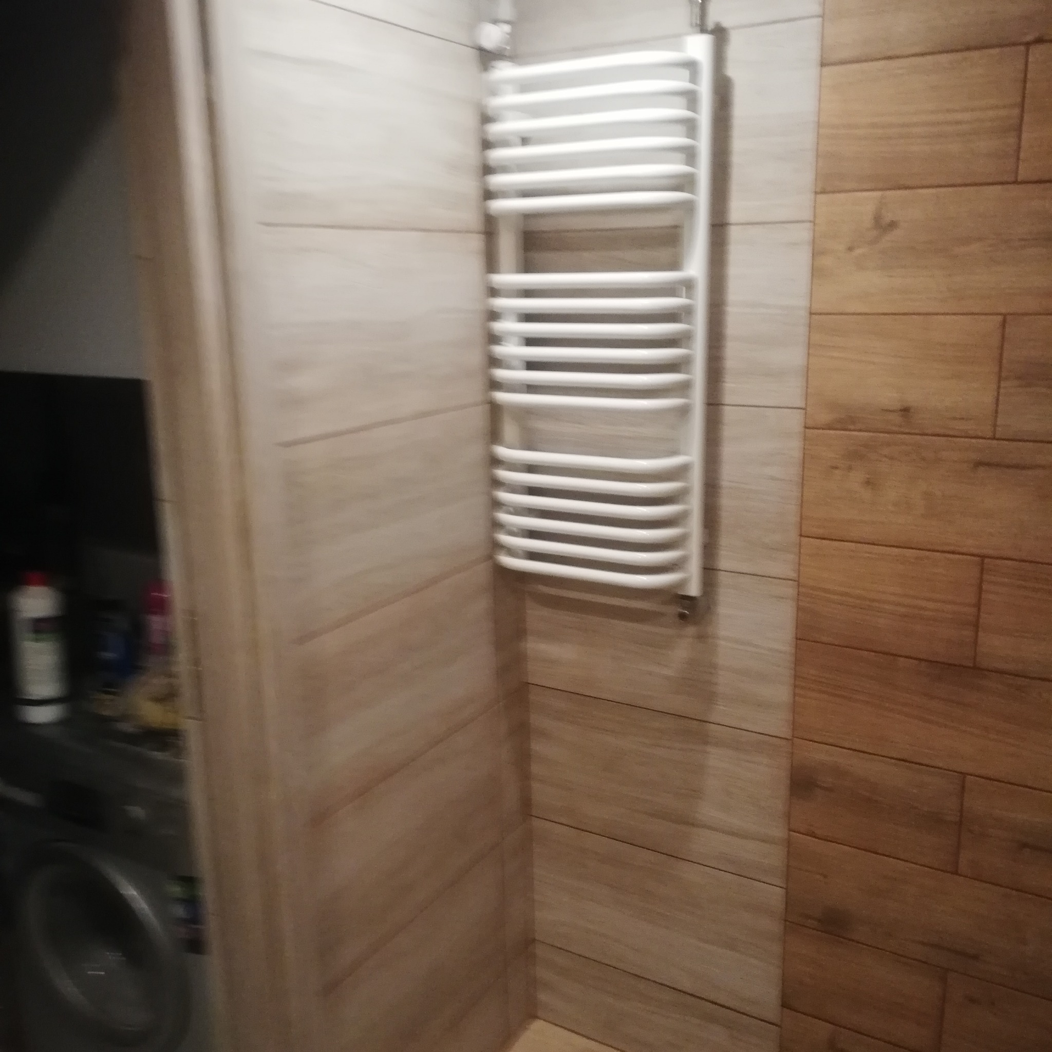 Biały, drabinkowy grzejnik łazienkowy zamontowany na ścianie wyłożonej płytkami imitującymi drewno, widoczna pralka i chemia gospodarcza w tle.