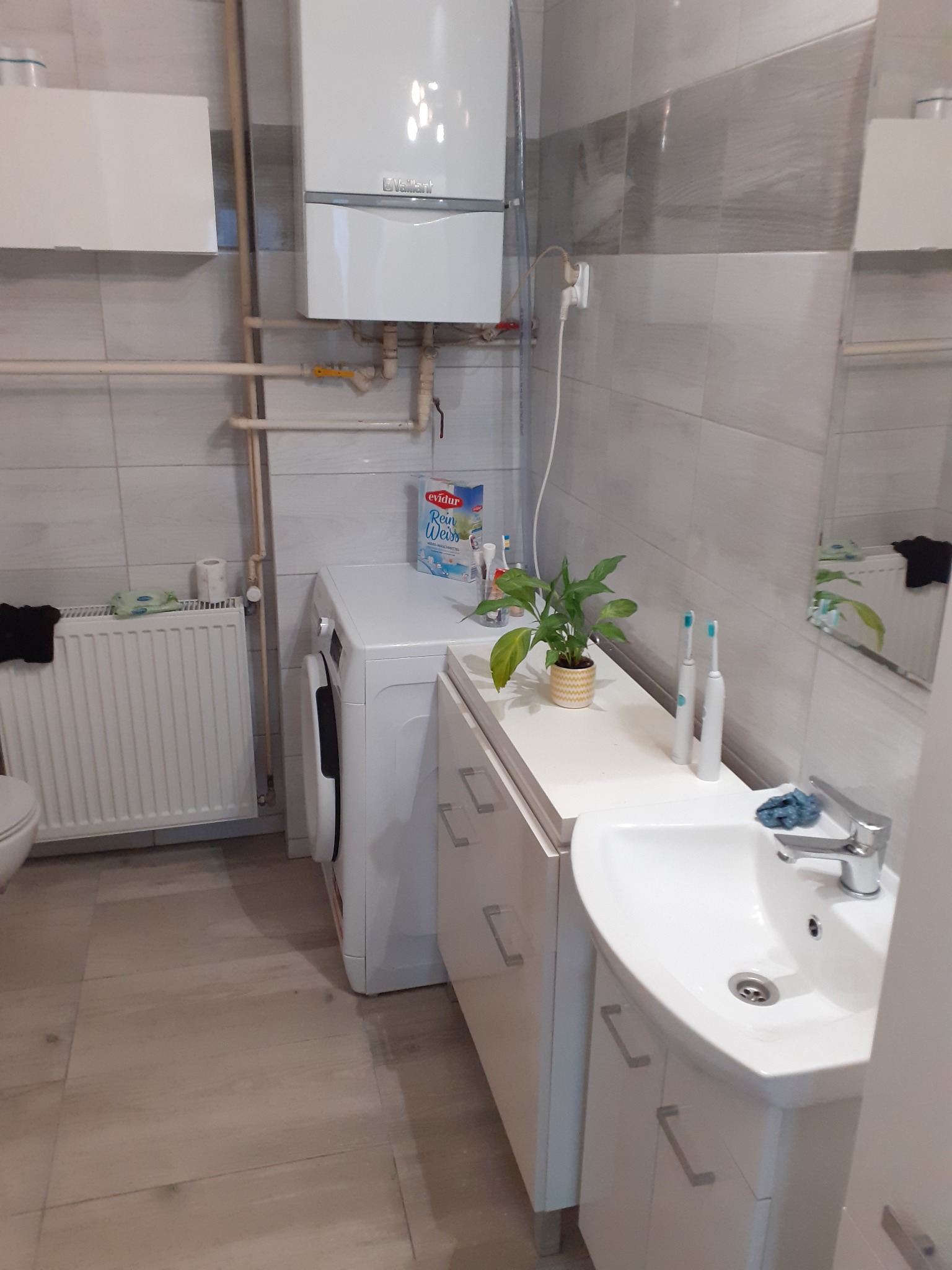 Funkcjonalna łazienka z szarymi płytkami, białym piecem gazowym, pralką i umywalką z szafką, uzupełniona o roślinę doniczkową i dwie szczoteczki elektryczne, z widocznym grzejnikiem i fragmentem...